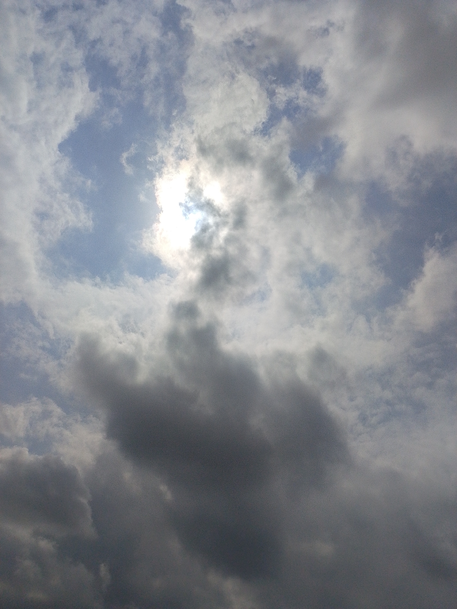 The image depicts a cloudy sky with the sun partially obscured by clouds, creating a soft, diffused light. The clouds vary in density and color, ranging from bright white to dark gray, indicating different atmospheric conditions. The sky is predominantly blue with patches of lighter blue peeking through the clouds. There are no discernable landmarks or objects in the foreground or background that would indicate a specific location within Lagos, Nigeria. The image is a close-up view of the sky, with no people, subjects, or actions visible.