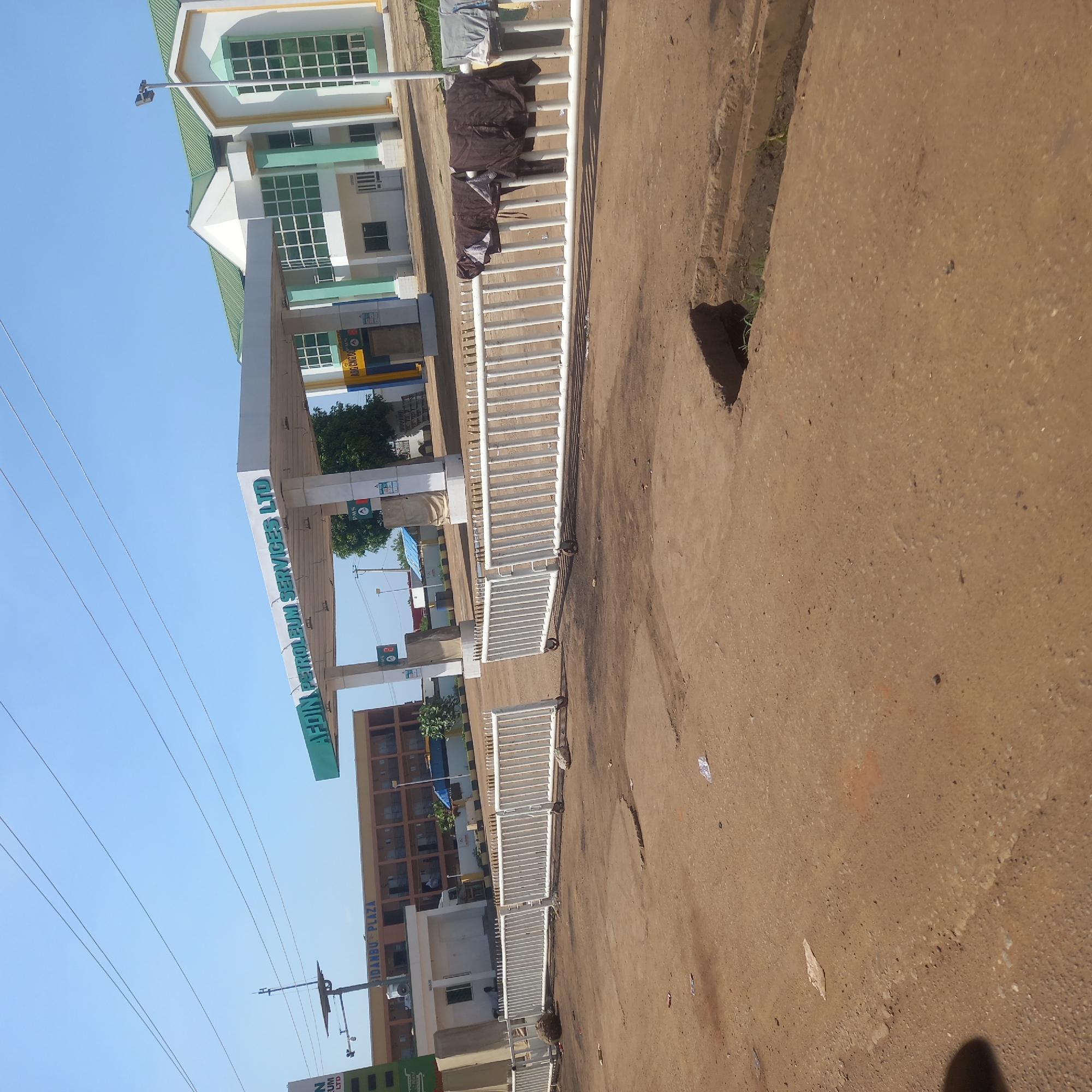 The image depicts a street scene in Hayin Banki, Nigeria. In the foreground, a dirt road with visible tire tracks and debris leads towards a white metal barricade. The barricade appears to be a portable gate or fence on wheels, with some clothing hanging on it. Beyond the barricade, a modern building with a green roof and white and light green facade stands prominently. A covered structure, likely a gas station canopy with signage, is visible in front of the building, bearing the text "AFDIN PETROLEUM SERVICES LTD". Several gas pumps are partially visible under the canopy. To the left, a multi-story building labeled "IDAMBU PLAZA" is visible. The sky is clear and blue. Power lines crisscross the upper portion of the image. The overall environment appears to be a commercial area. There are no people clearly visible in the frame.
