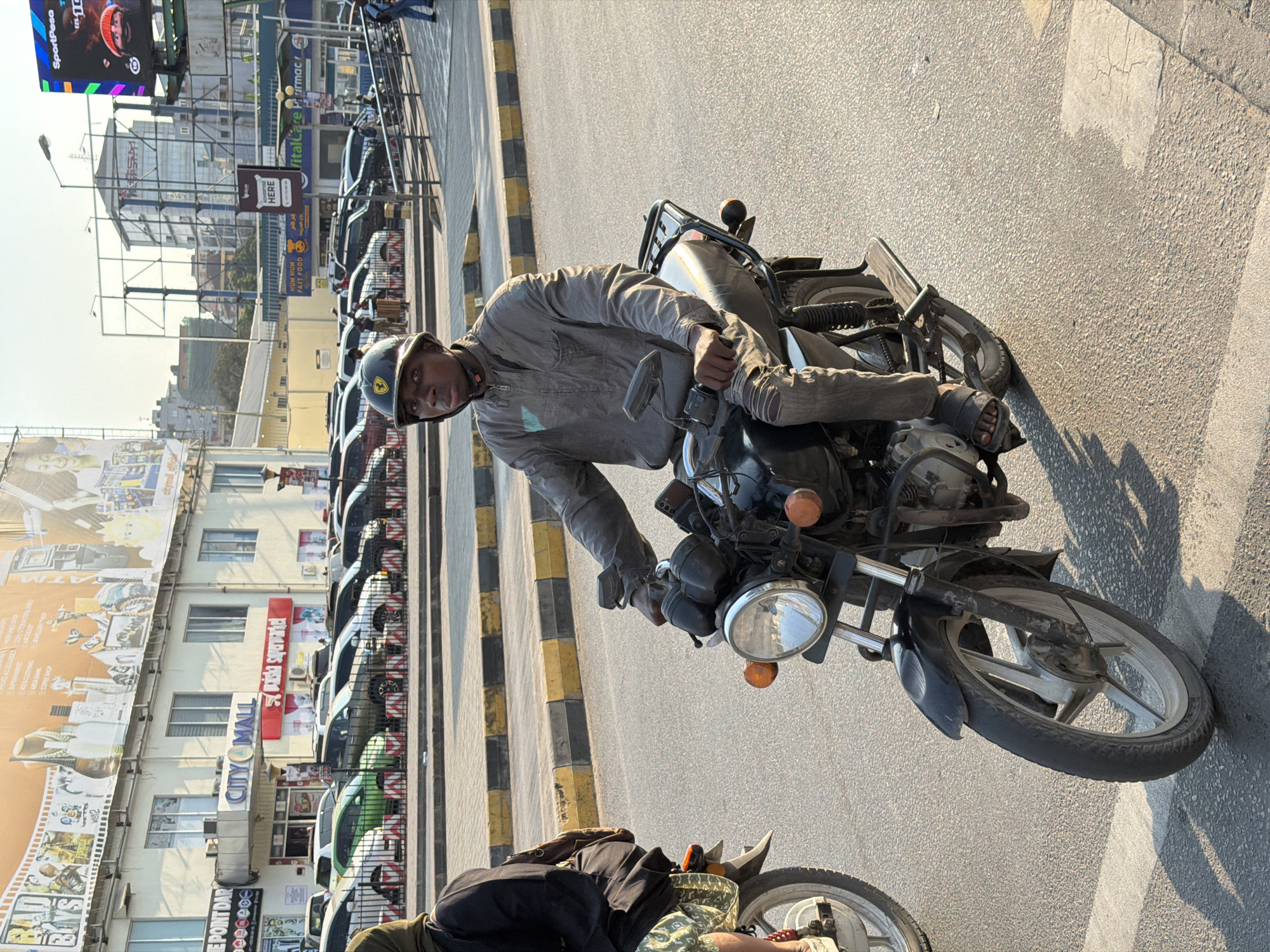 In Dar es Salaam, Tanzania, a man wearing a grey jacket, ripped jeans, and a black helmet rides a black motorcycle down a paved road. He is wearing dark sandals and is looking towards the camera. Another person on a motorcycle is visible in the background. The scene takes place in an urban environment with buildings and vehicles lining the street. The buildings include signage for "CITY MALL" and "SportPesa." The time of day appears to be late afternoon, with long shadows cast by the setting sun.