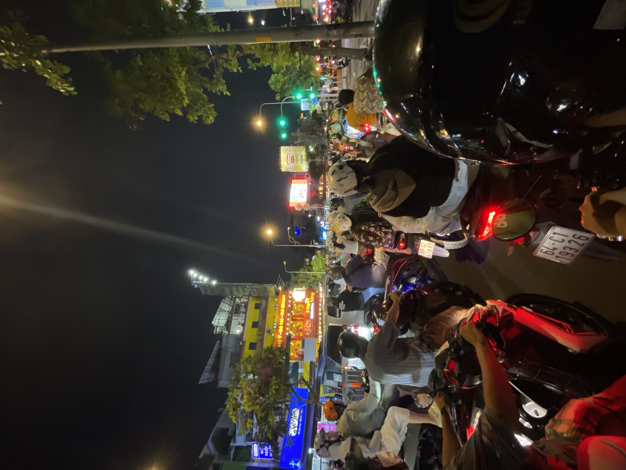 The image shows a crowded street in Thủ Đức, Vietnam, at night. Numerous motorbikes are waiting at a traffic light. Streetlights and illuminated billboards are visible in the background. A building with food advertisements, including "Mỳ Ý-Bò Né-Bó Bittet" and "3 Ngon Đô Nương Tảng," is present. License plates from the motorbikes are visible, including "59-G2 478.09" and "64-C1 193.26." People on motorbikes are wearing helmets. The traffic light is green.