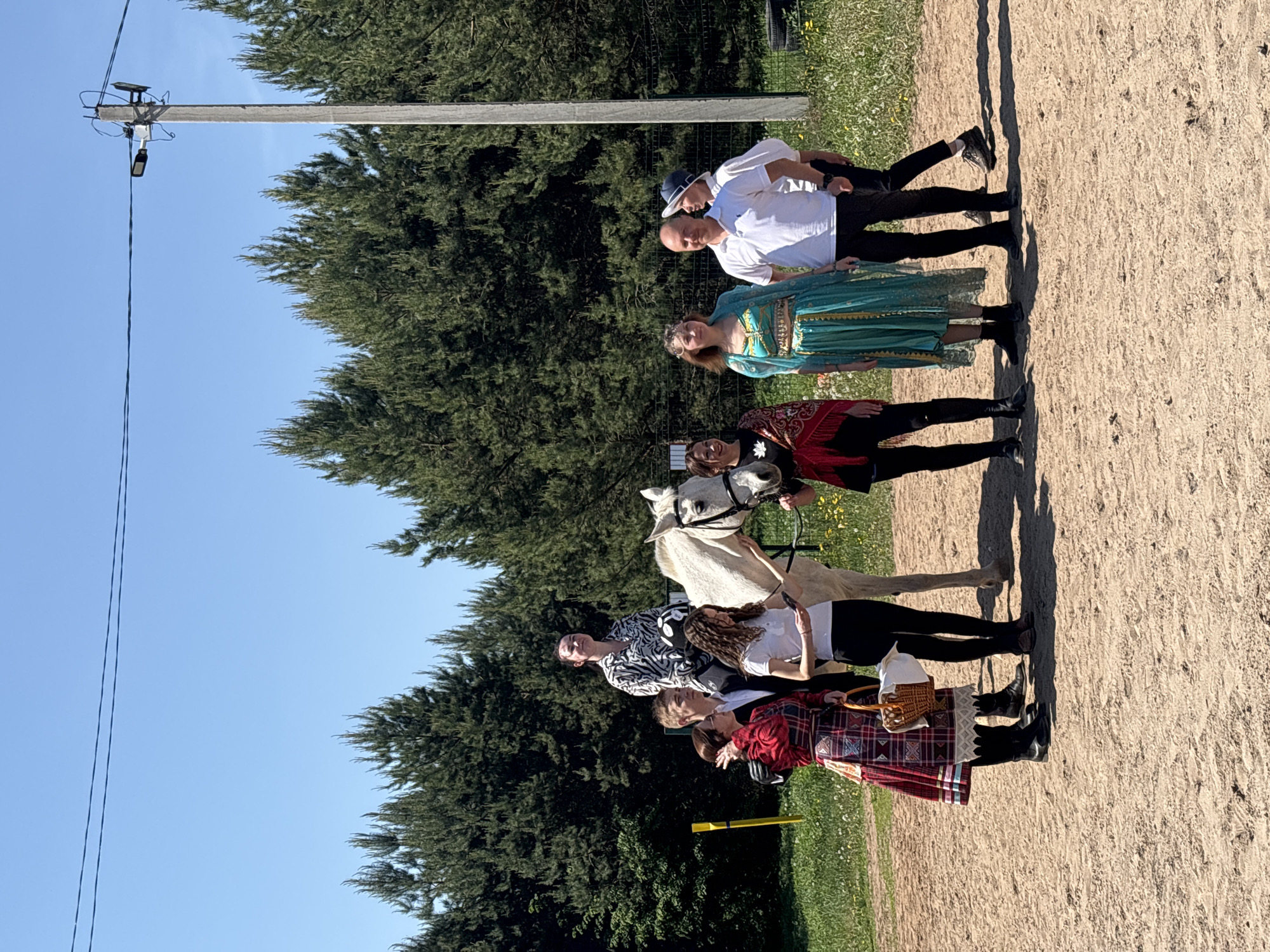 The image shows a line of six people, one on a horse, standing in a sandy area next to a grassy field with trees in the background under a clear blue sky. There is a utility pole visible. The group of people appear to be wearing costumes. The person furthest from the viewer is wearing a white shirt and dark pants, and the person next to them is wearing a green dress. The person after that is wearing a red and black dress. A white horse is standing between the third and fourth person. The fourth person is wearing a white shirt with a black pattern and black pants. The fifth person is wearing a white shirt and black pants. The last person closest to the viewer is wearing a red plaid skirt. Based on visual information, the location cannot be identified as Русский Вожой, Russia.