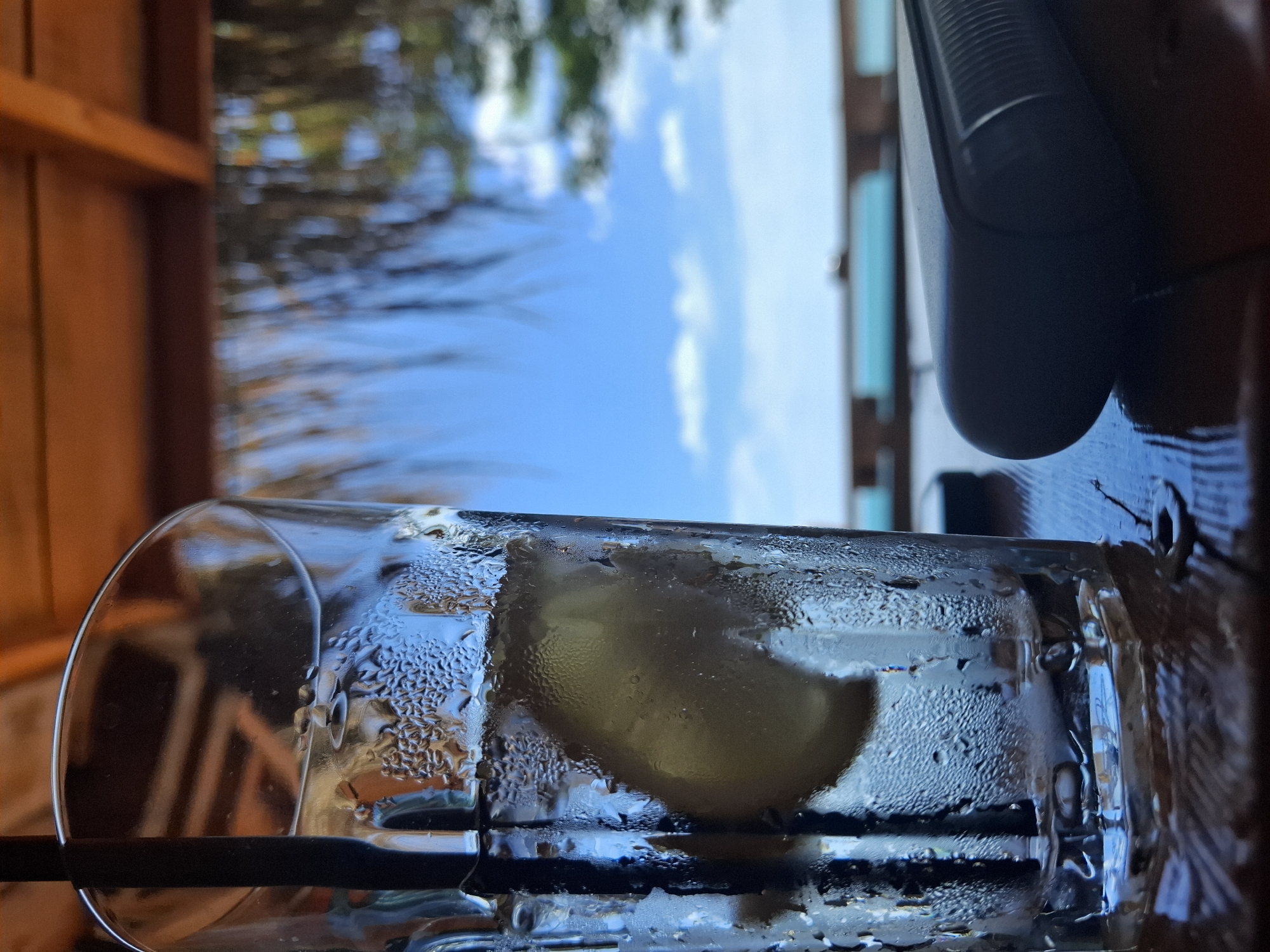 A close-up, low-angle shot shows a glass of liquid with condensation on the exterior, containing a lemon slice and a black straw. The glass sits on a dark, wooden surface. The background reveals a partly cloudy, blue sky, visible through what appears to be a partially open structure with wooden beams. The city is Tsarevo, Bulgaria, but no additional city landmarks are visible in the image.