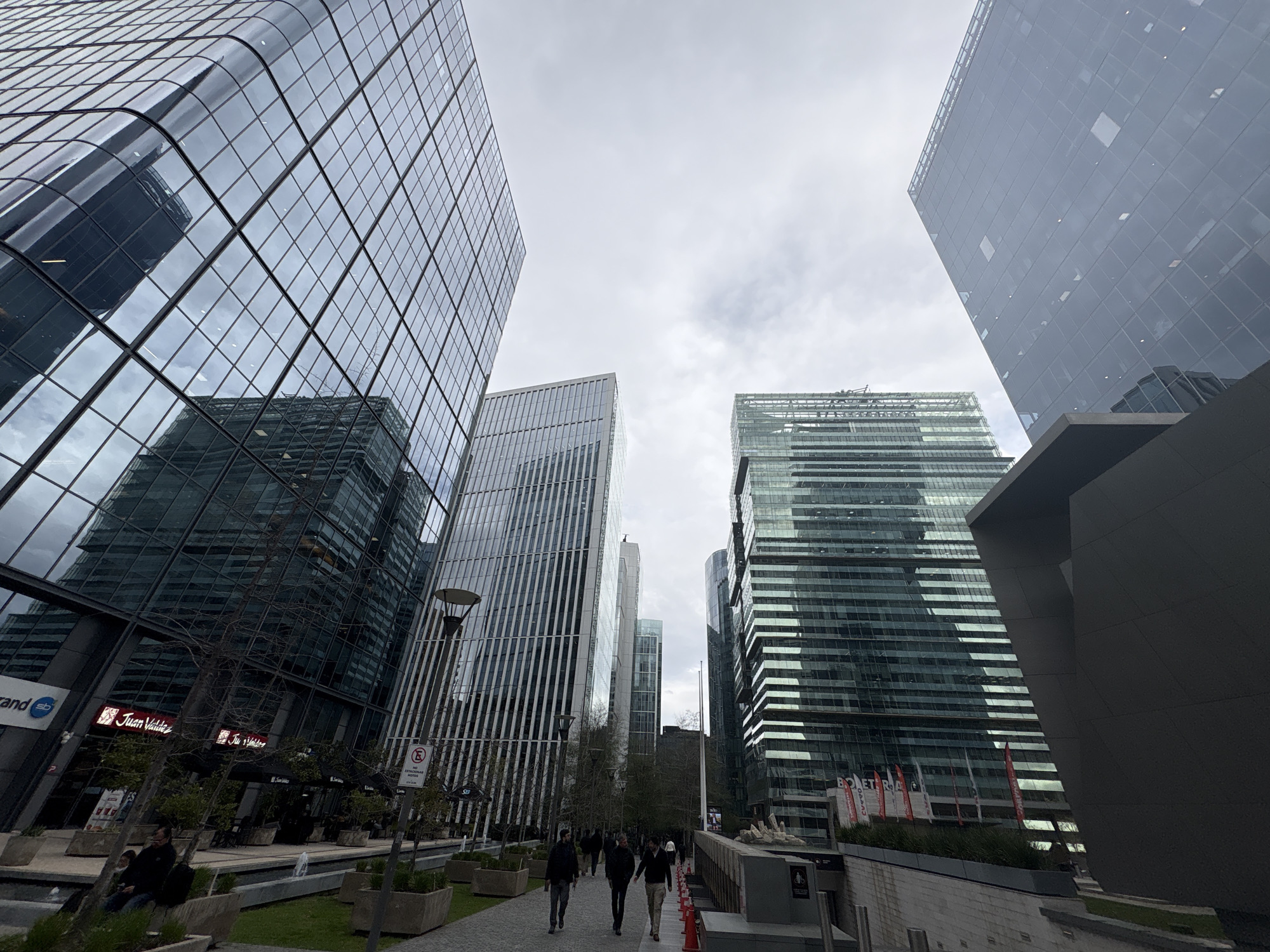 The image shows a wide-angle view of a modern urban plaza in Santiago, Chile, characterized by sleek, glass-covered skyscrapers. In the foreground, a paved walkway runs through an area with planters and benches. Several people are walking on the walkway, some dressed in casual outerwear. The buildings feature reflective glass facades, some with curved designs, and geometric patterns. One building prominently displays a sign for "Juan Valdez." Streetlights and trees are visible, contributing to the urban landscape. The sky is overcast, with clouds diffused across the frame. The perspective is low, looking upwards towards the towering structures.