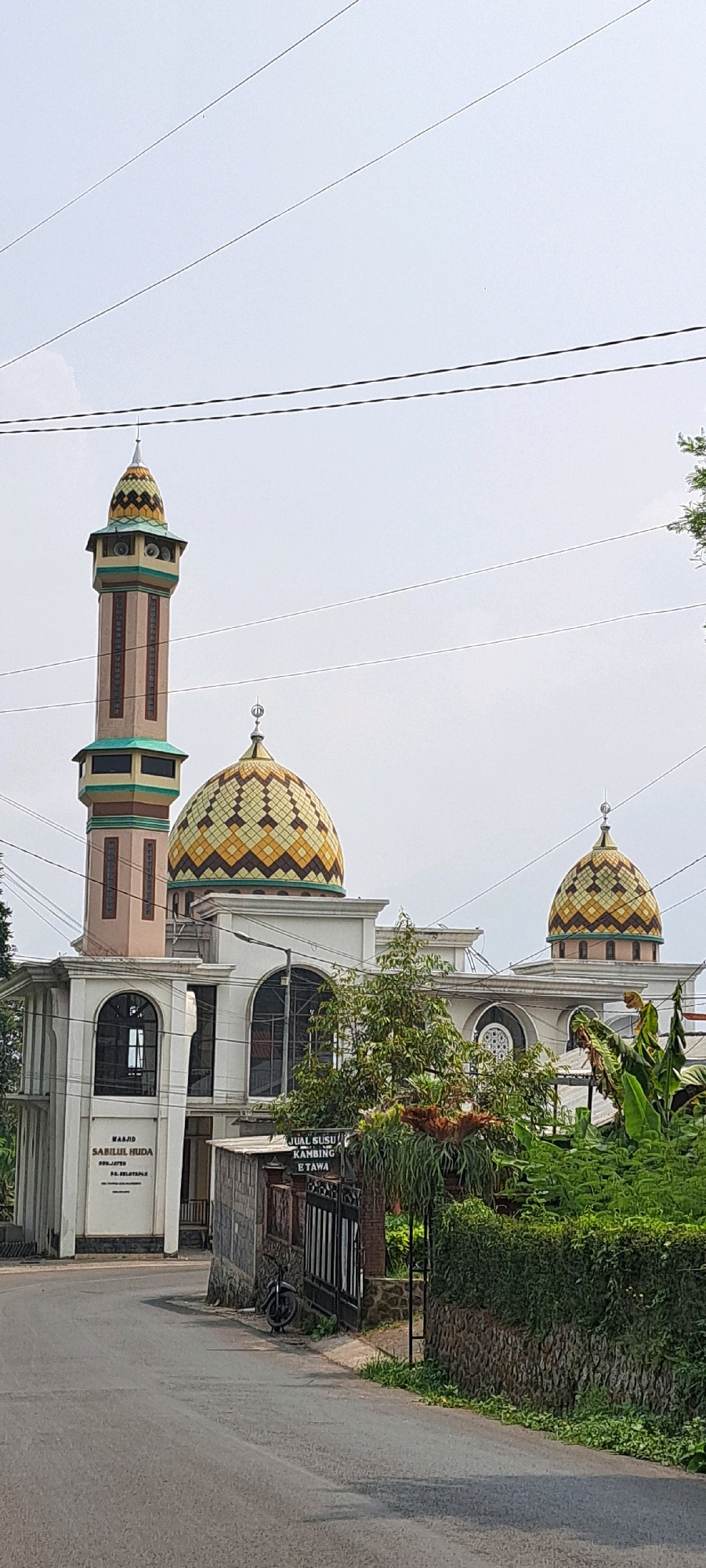 The image shows the Sabilul Huda Mosque in Selotapak, Indonesia. The mosque features a tall, cylindrical minaret with a conical roof and a patterned dome. The main building has a white facade with arched windows and another smaller patterned dome. In front of the mosque, there is a road, a fence, some greenery, and a parked motorcycle. A sign on the fence reads "JUAL SUSU KAMBING ETAWA," indicating the sale of goat milk. The sky is overcast.