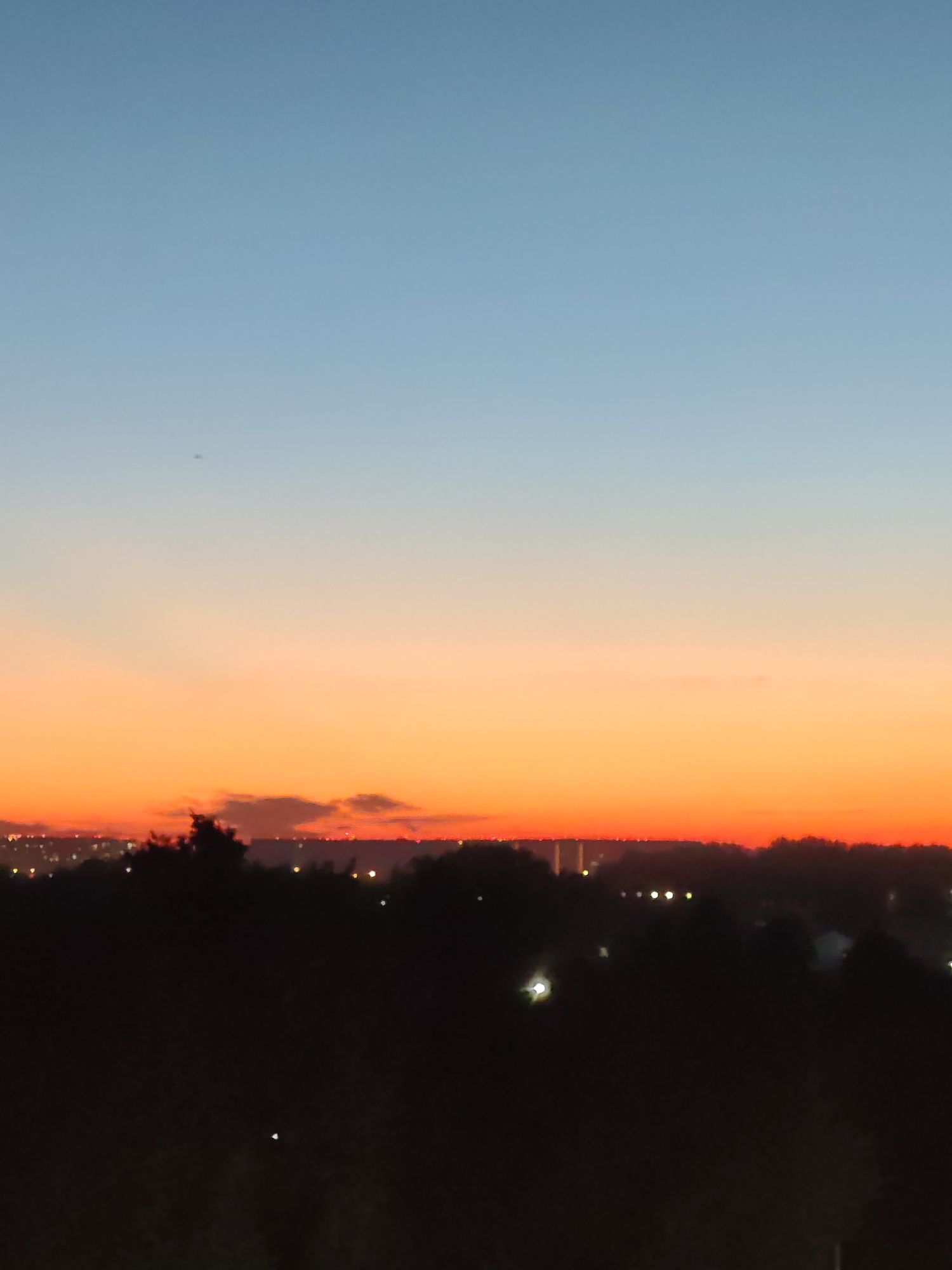 The image captures a twilight sky over a landscape that appears to be on the outskirts of Podolsk, Russia. The sky transitions from a pale blue at the top to a vibrant orange and pink gradient near the horizon, indicating either sunrise or sunset. Silhouetted against this colorful sky is a dark, undulating treeline, suggesting a natural or parkland area. Beyond the trees, a distant city skyline is faintly visible, marked by scattered lights, hinting at urban development. The overall impression is one of a peaceful, perhaps rural or semi-urban, setting during the transition between day and night.