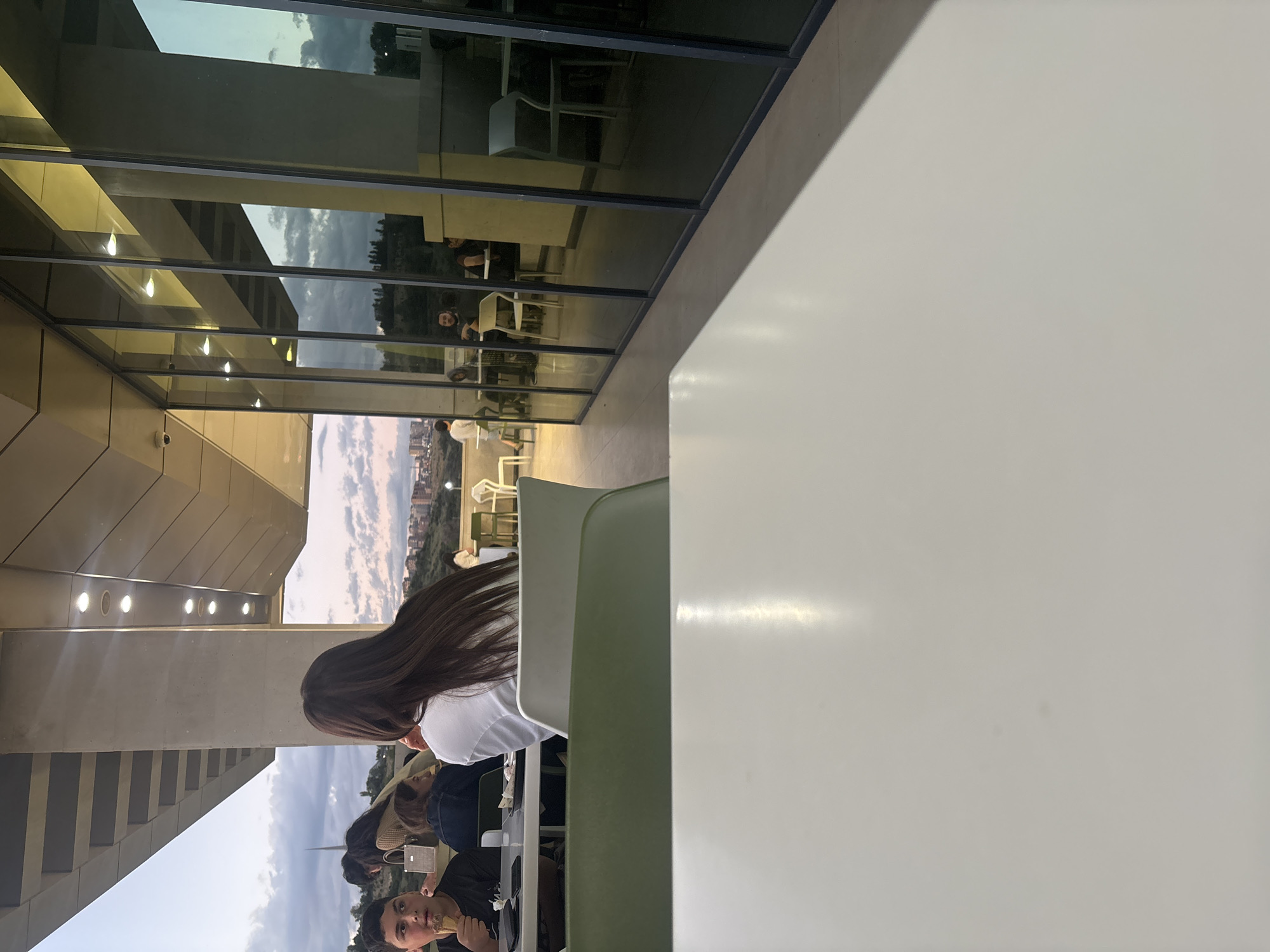 This image, originally oriented vertically, captures a scene inside what appears to be a modern cafe or dining area in Yerevan, Armenia. A large, plain white or light gray table occupies the right foreground. To the left, several individuals are seated at tables.
In the mid-ground, a person with long, dark hair, wearing a white top, is seen from behind, facing away from the camera. Below this person, a young male is visible in the lower-left, holding and eating an ice cream cone, looking directly towards the camera. Other partially obscured figures are also seated nearby. The seating area includes minimalist green and white chairs and tables.
The background is dominated by a large glass window or wall, offering a view and reflection of the exterior. The reflections show a partially cloudy sky and distant city buildings, indicating an urban setting. Inside, architectural elements such as concrete beams and an array of overhead recessed lights are visible. The interplay of natural light from outside and the activated interior lighting suggests it is late afternoon or early evening. The overall atmosphere appears casual, with people engaging in relaxed activities like eating and socializing. There is no discernible text visible in the image.