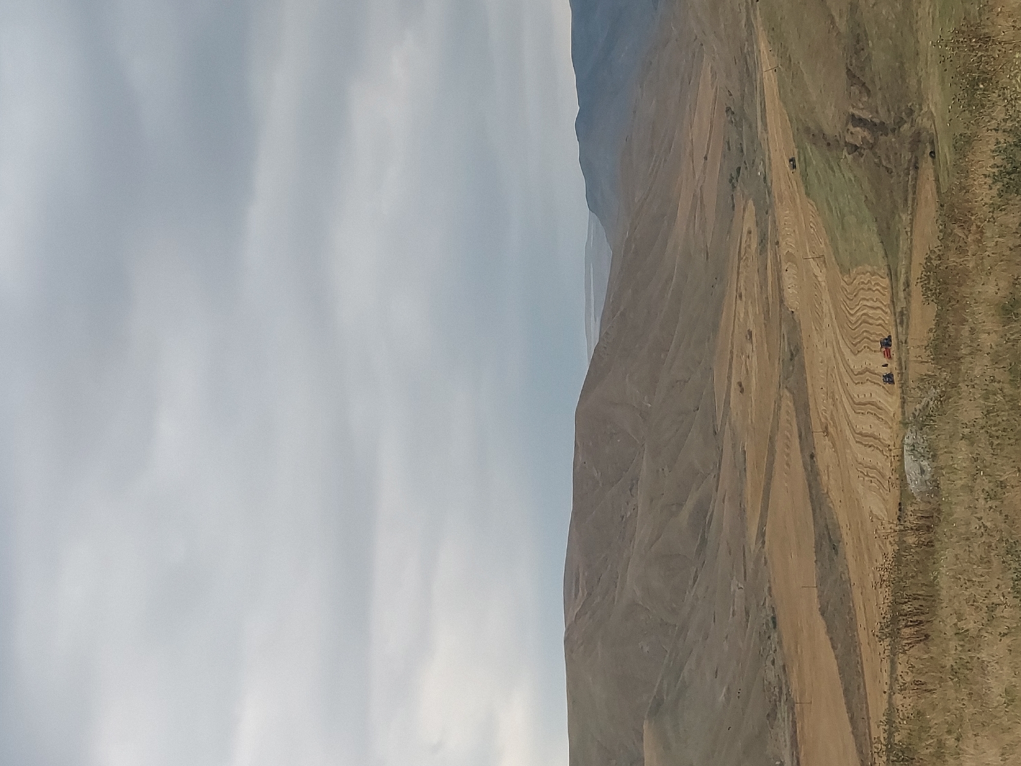 The image depicts a wide, panoramic view of a mountainous landscape. The scene is split vertically, with the left half dominated by a hazy, overcast sky, and the right half showcasing the arid, rugged terrain. The mountains are characterized by steep slopes and textured surfaces, with shades of brown, tan, and muted green indicating dry vegetation and earth.
In the middle ground of the right side, a dirt road or track winds through the landscape. Along this track, there are several small objects that appear to be vehicles. Two of these vehicles are distinctly red and blue, possibly flags or small structures, suggesting human presence or activity. Further up the slope, a dark-colored vehicle, possibly a tractor or truck, is visible. The terrain shows signs of human intervention, with visible tracks and cleared areas, hinting at agricultural or construction activities. The vegetation is sparse, consisting of dry grasses and scrub.
The lighting suggests it might be daytime, but the overcast sky creates a diffused light, with no strong shadows. The overall mood is one of vastness and natural, unpopulated beauty, with subtle indications of human interaction with the environment. The image is oriented vertically, giving the impression of looking down from a higher vantage point or across a very expansive vista. No visible text is present in the image.