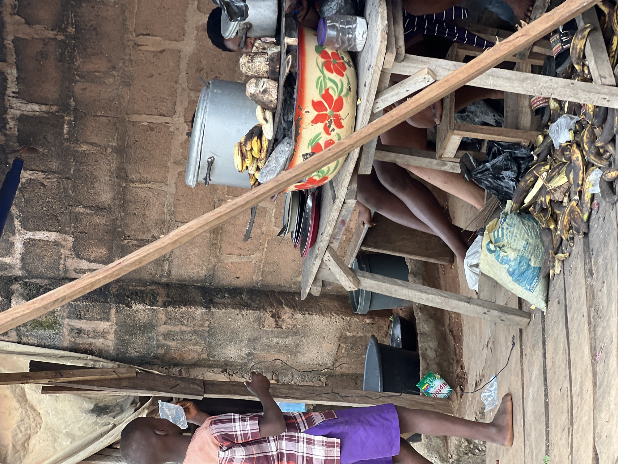 This image captures a scene at a market stall in Nkpor, Nigeria. A young boy stands on the left, wearing a plaid shirt and purple shorts, holding a plastic sachet and a string. In the background, a rough brick wall forms the backdrop. A wooden table is set up, laden with cooking utensils, including large pots, a decorative bowl filled with food, and various food items like plantains and yams. Several people, mostly their legs and torsos, are visible under the table and behind it, suggesting they are attending to the stall. The ground is wooden planking, strewn with discarded banana peels and other debris. Two large black plastic buckets are situated on the ground to the left of the stall. The overall impression is one of a busy, utilitarian market environment.