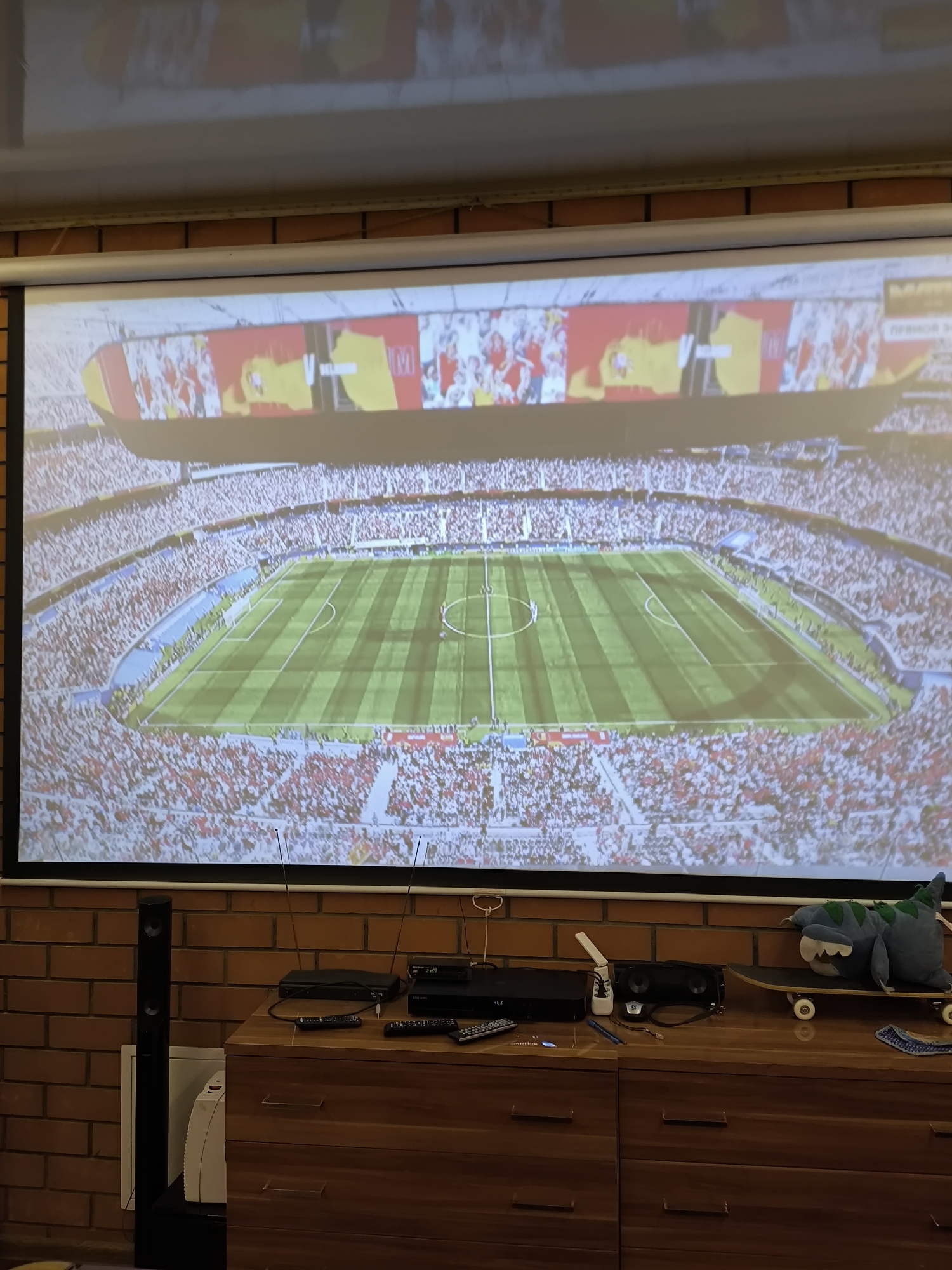 The image shows a projector screen displaying a soccer stadium filled with spectators. The stadium is vibrant with red and yellow banners visible in the stands. Below the screen, a wooden dresser with several drawers holds various electronic devices, including DVD players, a soundbar, and a remote control. A skateboard with a plush toy monster is placed on top of the dresser. To the left of the dresser, a tall black speaker stands next to a white heater. The background is a brick wall. The scene is likely a living room or entertainment area, and no specific location in Pokrov, Russia, can be identified.