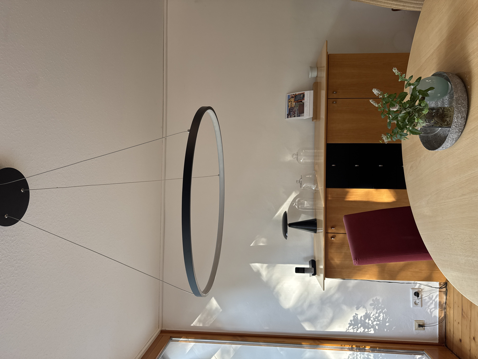 The image depicts a dining area in a residence in Rheine, Germany. A large, circular, modern pendant light fixture hangs from the ceiling over a light wood dining table. On the table are a cylindrical glass vase containing a sprig of white-flowered greenery and a small, oval, light blue glass vase, both resting on a textured grey oval tray.
Behind the table, a burgundy upholstered dining chair is partially visible. To the right of the chair is a light wood credenza with black drawer fronts and cabinet doors. On top of the credenza, from left to right, are a black telephone, a black cone-shaped lamp, two glass dome-covered displays (possibly for food or objects), and a desk calendar showing a colorful image of buildings and text. A small white container is also on the credenza.
To the left of the credenza, near a window with wooden framing, sunlight casts dappled shadows of foliage on the white wall. On the floor, two electrical outlets are visible, with one having a black power cord plugged into it and extending towards an unseen appliance. The floor is made of light wood planks. The overall environment suggests a well-lit, contemporary living space.
