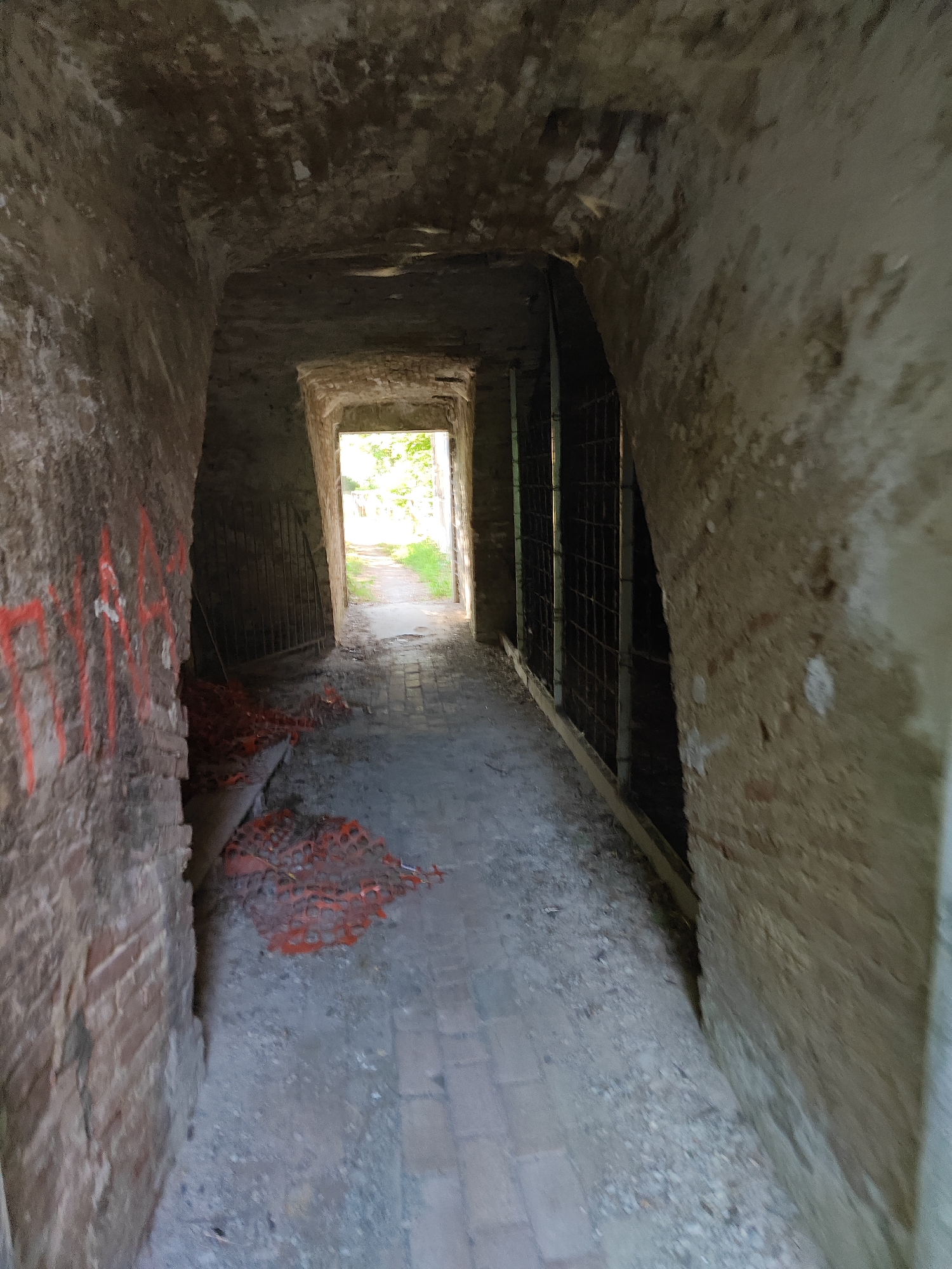 The image depicts a dimly lit, arched stone corridor in Modena, Italy. The walls are constructed of aged brick and rough stone, with some appearing to be partially rendered or plastered. There are areas of visible graffiti in red paint on the left-hand wall. The floor is paved with stones or bricks.
The corridor opens into a brighter area with a path leading outdoors, suggesting an exit or entrance. On the right side of the corridor, there are barred enclosures, reminiscent of windows or cell openings, made of metal grating. Orange construction netting or mesh is piled on the ground near the entrance of the corridor on the left. The overall impression is of an old, possibly abandoned or under-renovation, structure.