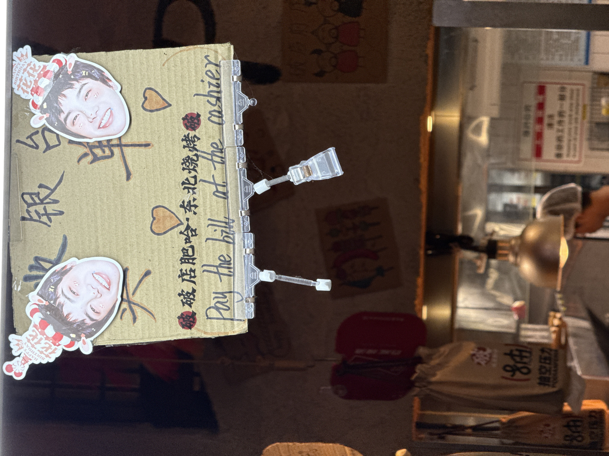 The image shows a street-level food stall or restaurant in Nanshan District, China. In the foreground, a cardboard sign is displayed, featuring handwritten Chinese characters and English text. The sign has two cutout pictures of a young person wearing a birthday hat, and the English text reads "Pay the bill at the cashier". Below the sign, two clear plastic clips are attached. In the background, blurry elements suggest the interior of a food establishment, including kitchen equipment like a stainless steel hood and possibly food items in bags. The lighting suggests an indoor or covered outdoor setting.
