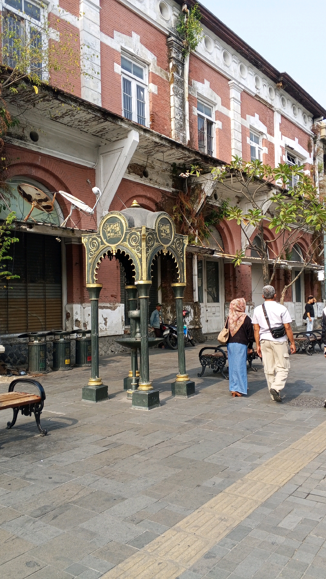 The image captures a street scene in Semarang, Indonesia, likely near the old city center, characterized by colonial-era architecture and a cobblestone street. In the foreground, a decorative black and gold fountain structure with ornate pillars stands prominently. Two individuals are walking away from the camera on the right side of the frame. The person on the left wears a hijab and a blue skirt, while the person on the right is a male in a white shirt and beige pants, carrying a black bag across his shoulder. A park bench with a wooden seat and a black metal frame is visible on the far left.
The background features a two-story brick building with white architectural accents, classic windows, and a weathered balcony. Vegetation is growing on the building's facade and balcony, suggesting age and neglect. The street is paved with stone tiles, and a tactile paving strip runs along the right side of the walkway, indicating accessibility features. Several dark green trash bins are positioned against the building on the left. A man is seated on a motorcycle near the building on the left, and another person is visible further down the street on the right. The lighting suggests it is daytime, with shadows indicating the sun's position.