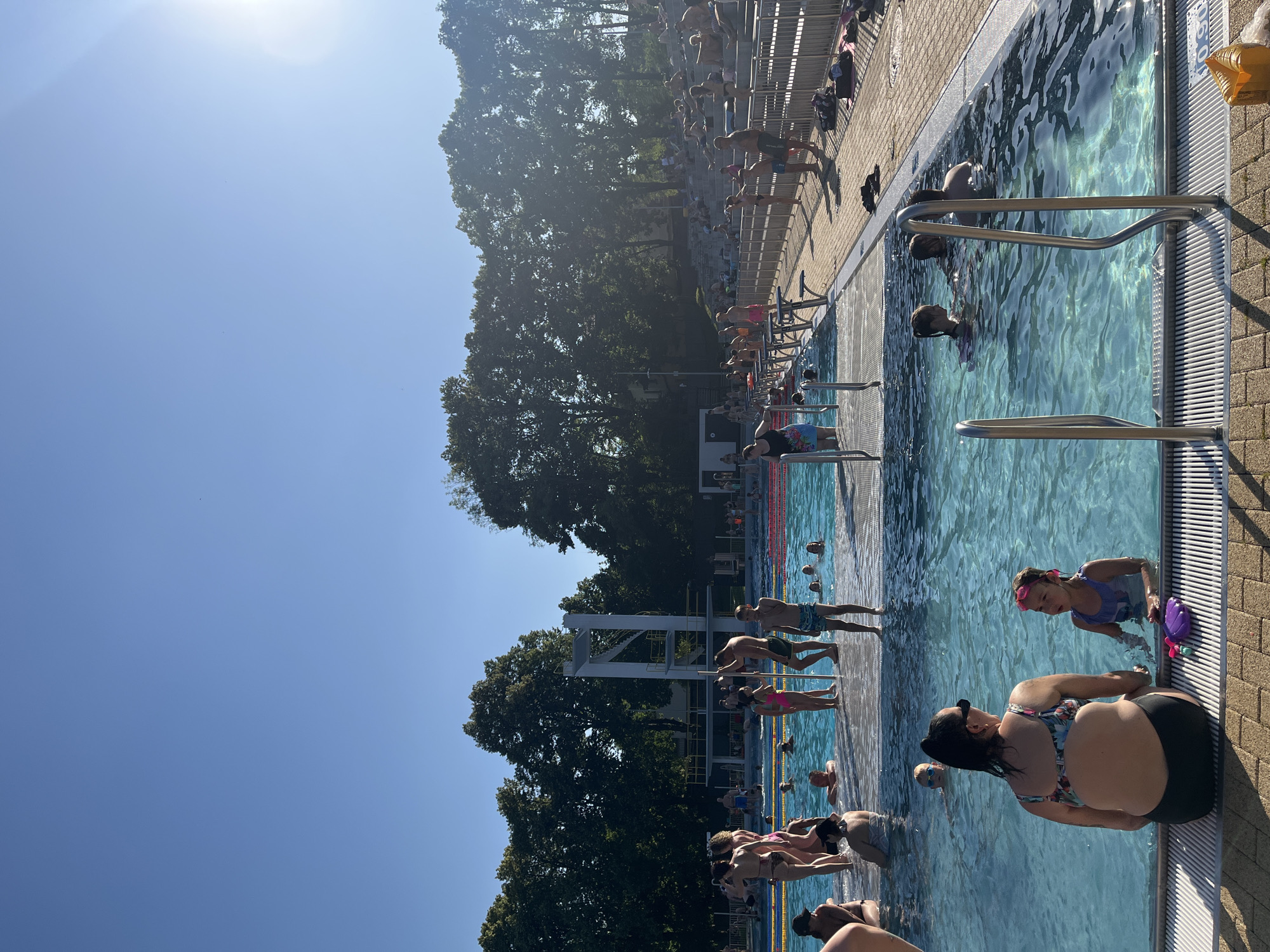 The image depicts a sunny day at an outdoor swimming pool, likely during the summer. The scene is filled with people of various ages enjoying the water and the surrounding area. In the foreground, a woman with dark hair and sunglasses is sitting by the pool's edge, looking towards the water. To her right, a young girl with pink goggles is in the pool, interacting with a purple pool toy. Further down the pool, several individuals are standing on starting blocks, seemingly preparing for a race or dive. Other people are visible in the water, some swimming, others wading. The pool area is lined with brick paving, and metal railings are present for swimmers to hold onto. In the background, lush green trees and a clear blue sky suggest a pleasant weather. The sun is high in the sky, casting shadows and indicating it is likely midday. There is no visible text in the image.