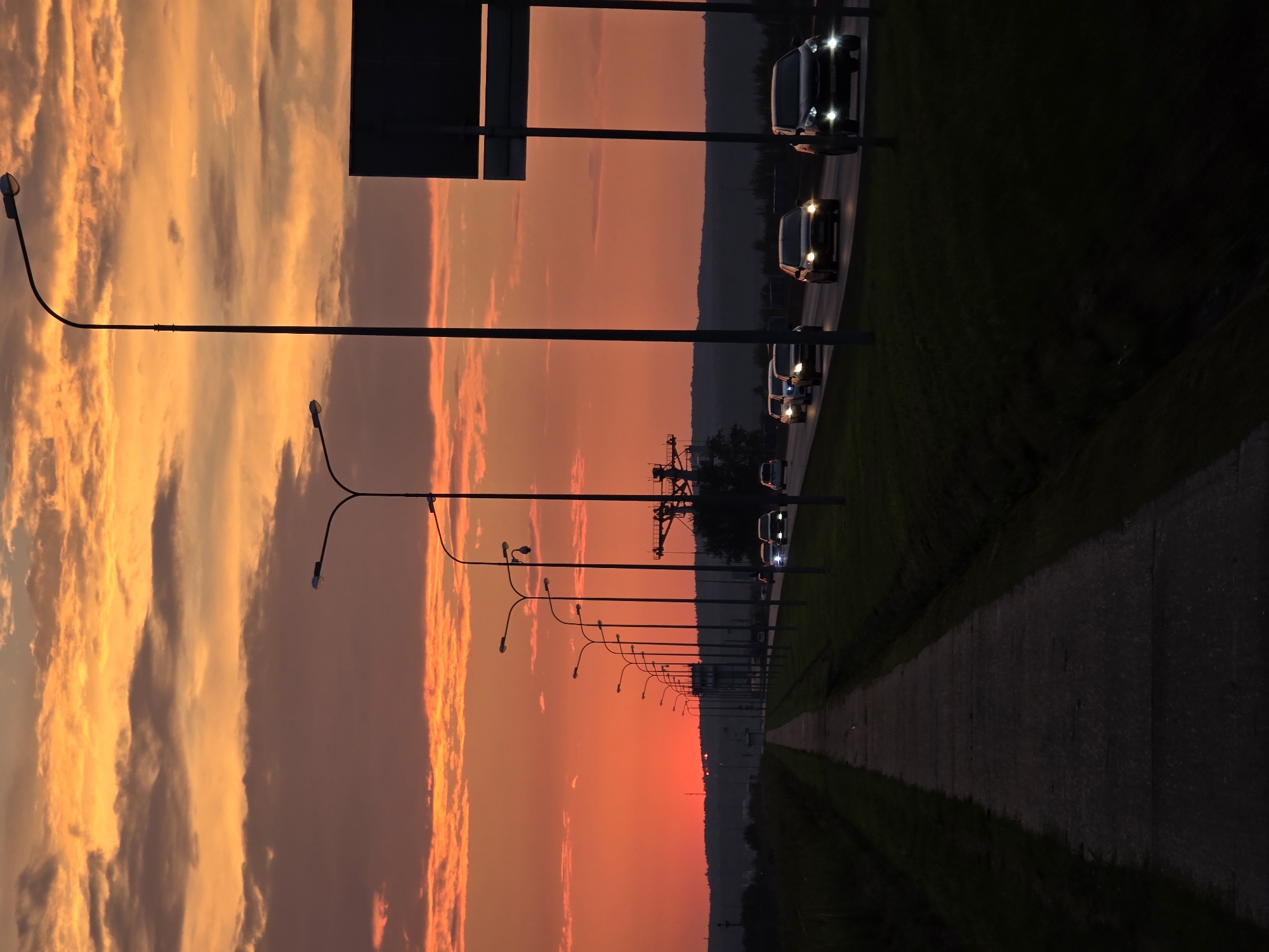 The image depicts a sunset scene with a fiery orange sky filled with scattered clouds over a semi-rural landscape. A paved walkway runs along the left side of the frame, disappearing into the distance. To the right of the walkway is a grassy verge and then a multi-lane road. Several cars are driving on the road, their headlights illuminated. A row of tall streetlights lines the side of the road. In the background, there is a dark, silhouetted treeline and hills. A large, dark rectangular sign is visible on a pole on the far right. The setting appears to be on the outskirts of Chaikovsky, Russia, possibly along a highway or main road leading into or out of the city. There are no people visible in the image.