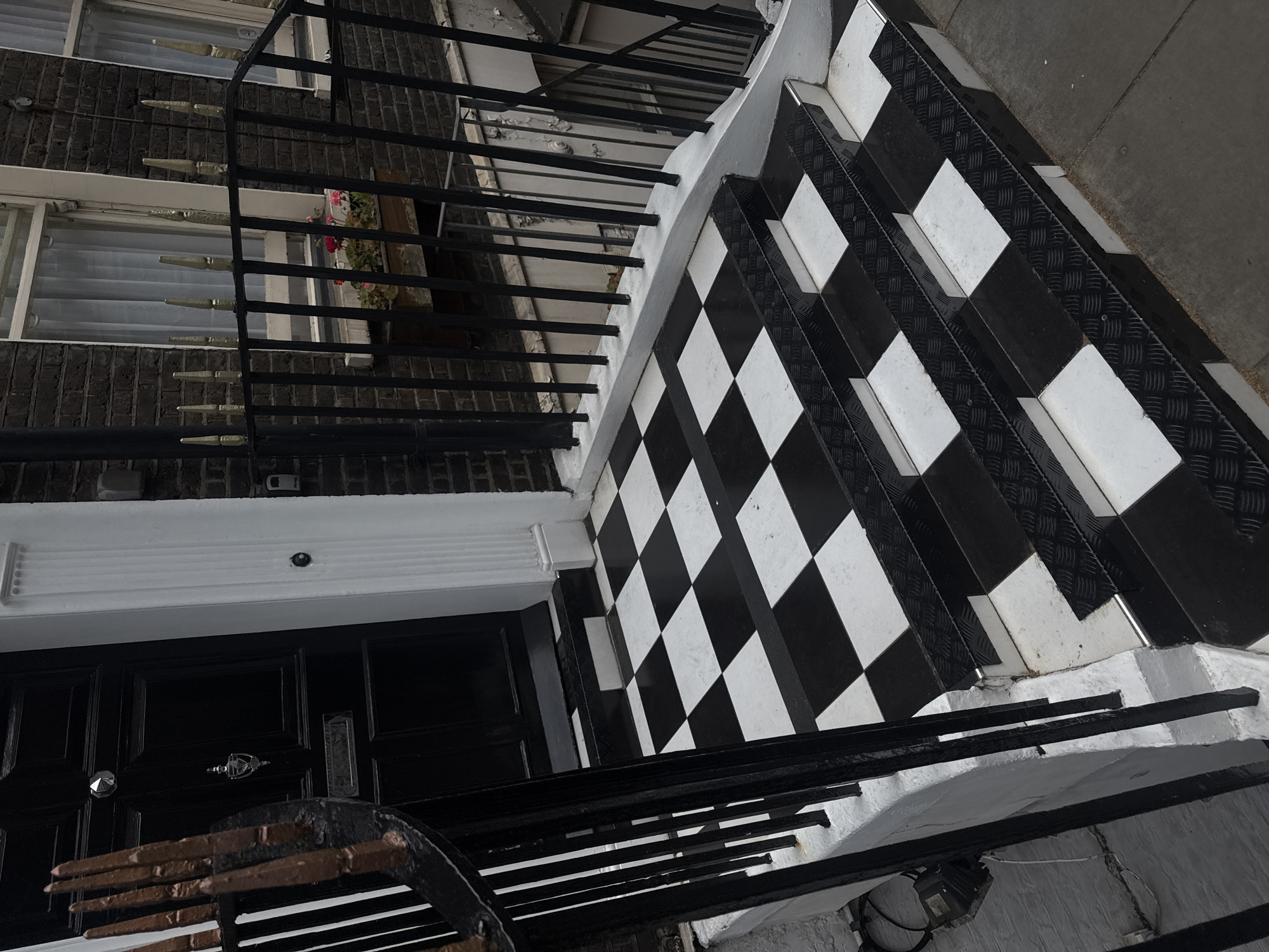 The image captures the entrance of a building, likely a townhouse or apartment block, located in London, United Kingdom. The scene is dominated by a striking black and white checkerboard tiled pathway and steps, leading up to a black front door.
The stairs feature black metal anti-slip plates on each tread, with alternating black and white tiles on the risers, creating a bold, geometric pattern. The tiled surface extends to a landing area in front of the door. A black iron railing, adorned with pointed, possibly brass-colored decorative finials, runs along the left side of the steps and landing, providing a safety barrier. Beneath the railing, the structural base is painted white, showing some wear and exposed wiring in places.
The building's facade is constructed from dark brick. Multiple windows with white frames and light, sheer curtains are visible on the upper levels. A window box overflowing with pink or red flowers can be seen under one of the windows, adding a splash of color to the otherwise monochrome palette.
The double front door is painted black and features silver hardware, including a door knocker, handles, and a letterbox. Above the door, a prominent white decorative molding or lintel is present. Small utility fixtures are mounted on the wall above the door and between windows.
No people are visible, and no specific activity is occurring. The lighting suggests daytime, possibly overcast or early morning/late afternoon, as there are no strong shadows, giving the scene a quiet and composed ambiance. No visible text is present within the image.