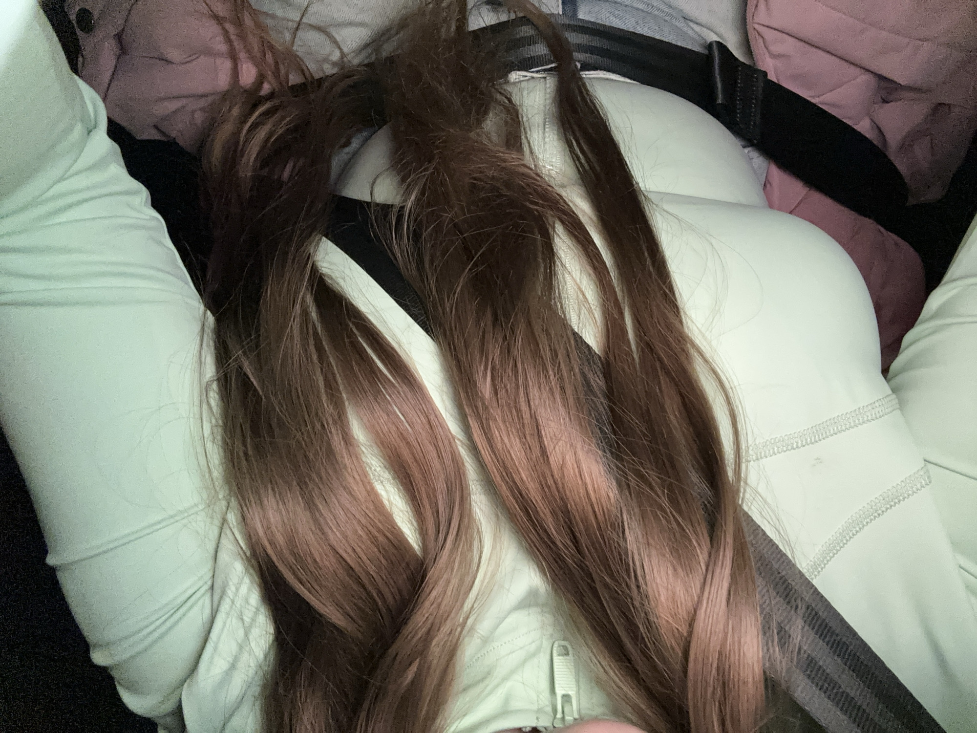 The image shows a person in a seated position, wearing a light green top. A black seatbelt is fastened across their body. Strands of long, brown hair are visible, partially obscuring the clothing. There is a zipper pull visible on the top. In the background, there is a glimpse of pink fabric and a blue denim material. The location is not discernible and there is no evidence it is Moscow, Russia.