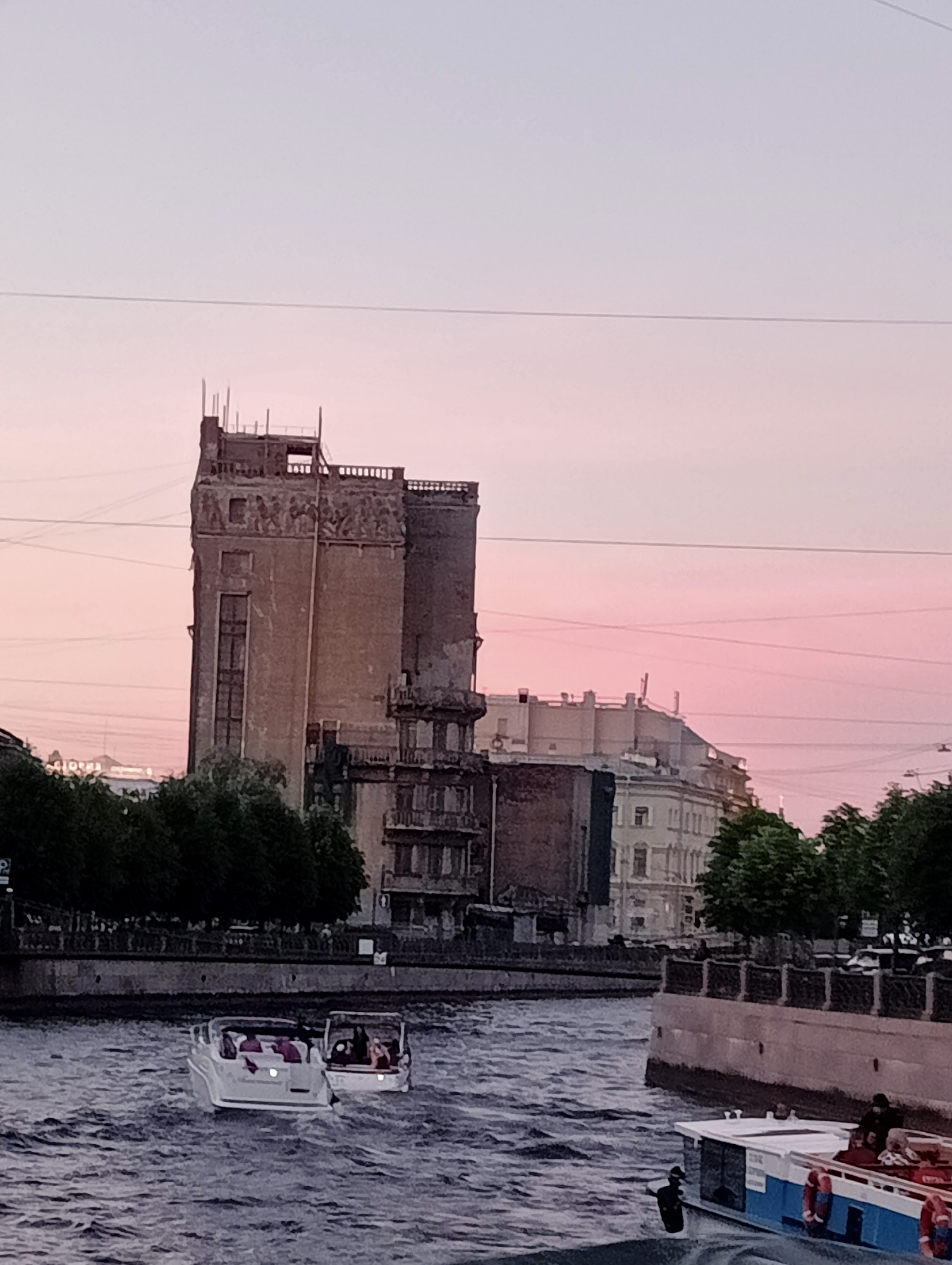 The image captures a view of the Neva River in Saint Petersburg, Russia, during what appears to be twilight, with a soft pink and lavender sky. Dominating the background is the imposing structure of the Lighthouse Building (Mayak Building), a historical industrial building that has been repurposed. Its weathered facade and varied architectural elements, including what seems to be remnants of decorative carvings and exposed upper-level construction, are visible. Adjacent to it, other buildings with neoclassical architectural styles line the riverbank, partially obscured by lush green trees.
In the foreground, two white motorboats are navigating the river. The boat on the left is further away and smaller, while the one on the right is closer and larger, with several passengers visible on deck. A second, larger boat with a blue stripe along its side is also present, docked or moving slowly in the lower right quadrant of the image, with a few individuals on its deck and one standing near its edge. The river's surface is rippled, indicating moderate movement. A pedestrian embankment with decorative railings runs along the river, with silhouetted figures of people visible strolling along it. Overhead, thin wires, likely for utilities, crisscross the sky.