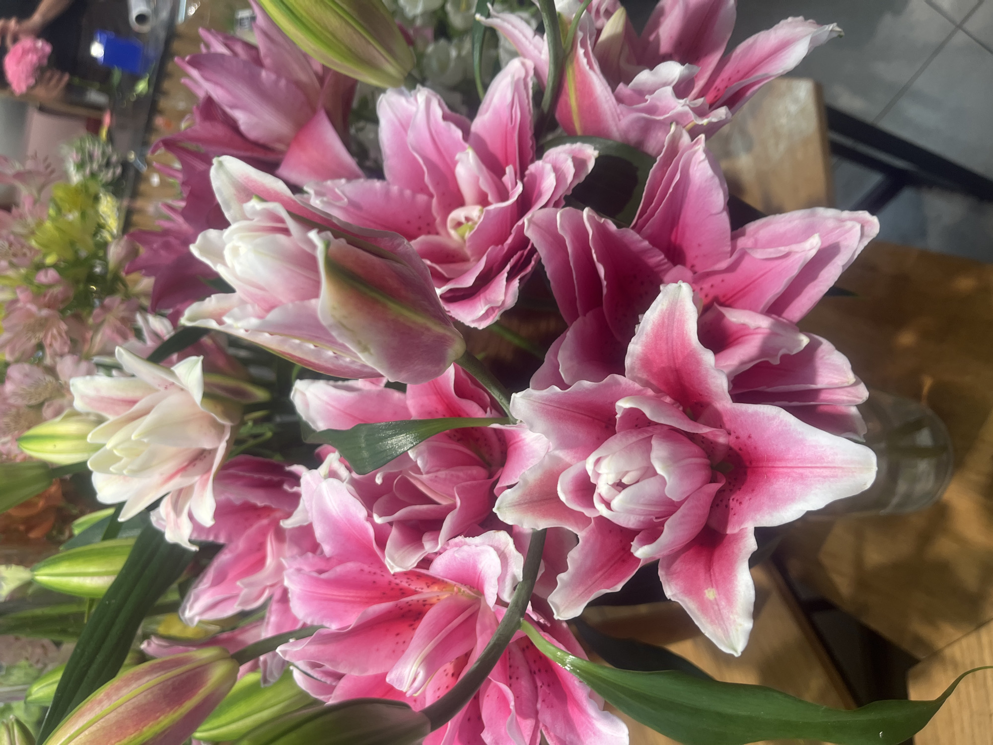 A close-up, eye-level shot shows a vibrant bouquet of pink and white lilies. The lilies are in various stages of bloom, with some fully opened and others still in bud. Their petals are soft and velvety, with delicate streaks of deeper pink and white edges. The leaves are a lush green, adding a contrasting pop of color. The bouquet is in a clear glass vase, which sits on a wooden surface. In the background, out of focus, is a blurred image of a retail environment, possibly a flower shop, with hints of other floral arrangements and shelving. No people are clearly visible. The lighting is natural and soft, highlighting the delicate beauty of the flowers. The image does not provide any specific information about its location in Đà Nẵng, Vietnam.