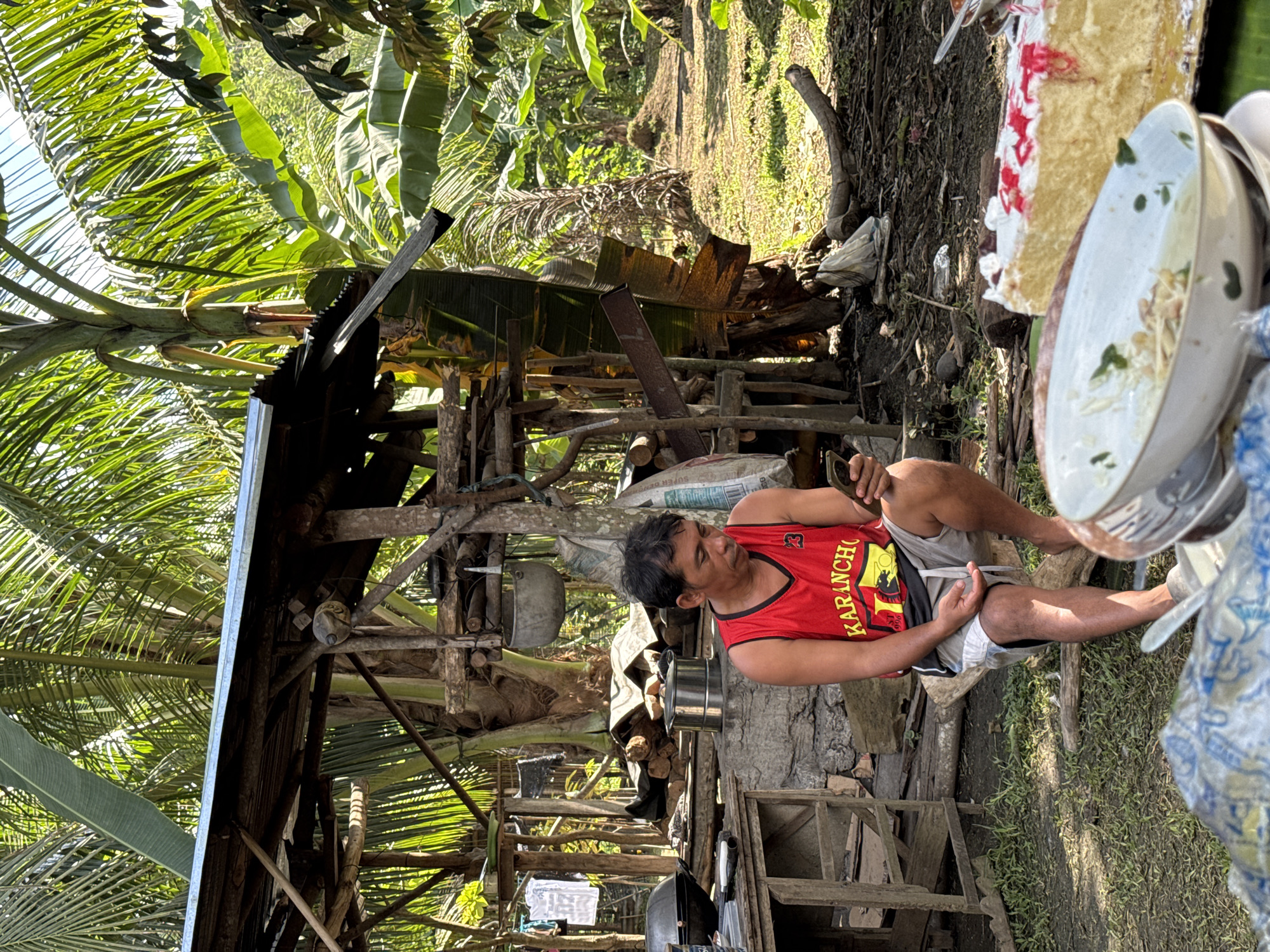 The image depicts an outdoor scene in Carmen, Philippines, under bright daylight. In the midground, an adult male, of apparent Asian ethnicity, is seated on a wooden log, clad in a red and black "KARANCHO 23" basketball jersey and light-colored shorts, operating a smartphone with both hands. His bare feet are visible on the ground. To his left, a rudimentary outdoor kitchen area is visible, featuring a wooden table, a large dark cooking pot on a stand, and a shiny metallic multi-tiered steamer pot atop a stone or concrete base. A white garment hangs behind this area. Various sacks and discarded items are scattered around the base of the structure.
The background is dominated by dense tropical vegetation, including large palm trees with fronds and broad-leafed banana trees, through which sunlight filters. A rustic, elevated wooden structure with a corrugated metal roof stands behind the man. Further back, a cleared patch of ground or dirt path is visible, bordered by more trees. The foreground, out of focus, contains a stack of dirty dishes, including a white bowl with food remnants, and a partially eaten cake with white frosting and red decoration on a gold board. Blue and white patterned fabric or plastic is also present in the immediate foreground. The ground is a mix of dirt, sparse grass, and organic debris.