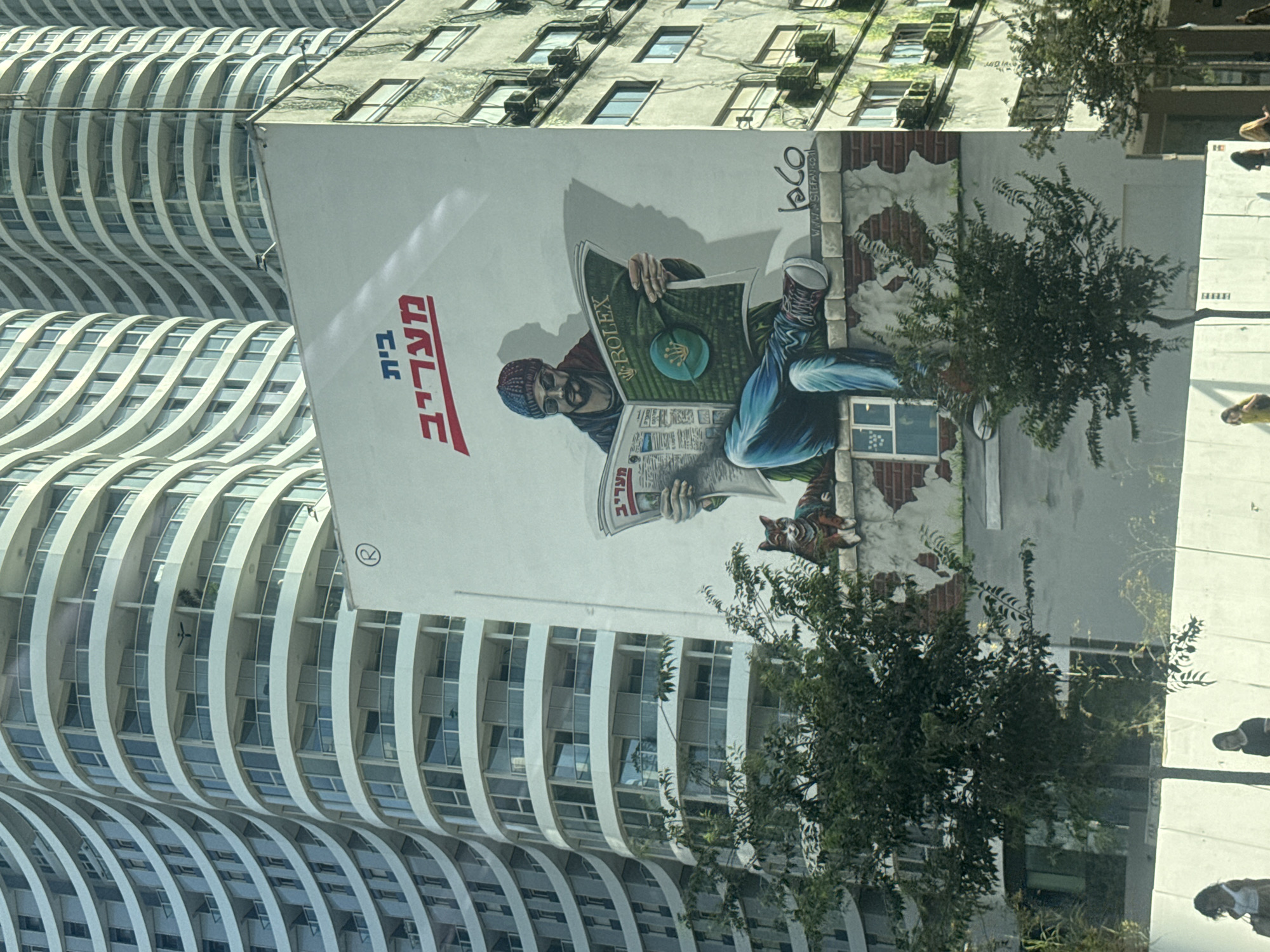 This image depicts a mural on the side of a building in Tel Aviv, Israel, with two modern, curved residential towers in the background. The mural features a man wearing a beanie, sunglasses, and casual clothing, sitting and reading a newspaper. He is depicted with his legs crossed, and he appears to be on a ledge. Next to him, a cat is also sitting, looking towards the viewer. The newspaper the man is reading has a prominent "ROLEX" logo on its front page, which is green and features the Rolex crown emblem. Above the mural, text in Hebrew reads "בית מעריב" (Beit Maariv). The building on which the mural is painted appears older than the surrounding towers, with visible signs of wear and air conditioning units on its facade. In the foreground, some trees and a low white wall with people walking behind it are visible, suggesting this is a street-level view.