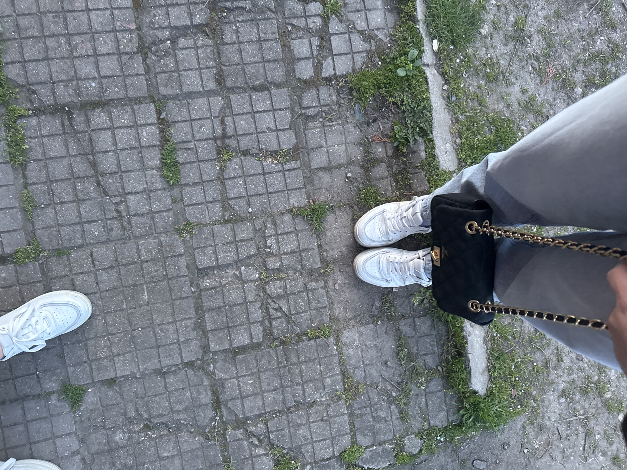The image shows a ground-level perspective of a person standing on a paved surface, likely a sidewalk or pedestrian area. The paving is made of square concrete tiles with patches of grass and moss growing in the cracks. The person is wearing light-colored jeans and white sneakers. A black handbag with a gold chain strap is positioned near their right leg. The overall environment appears to be an outdoor setting, potentially in an urban or suburban area. The location mentioned, Lehchevo, Bulgaria, suggests this is where the image was taken. No other people or interactions are visible in the frame.