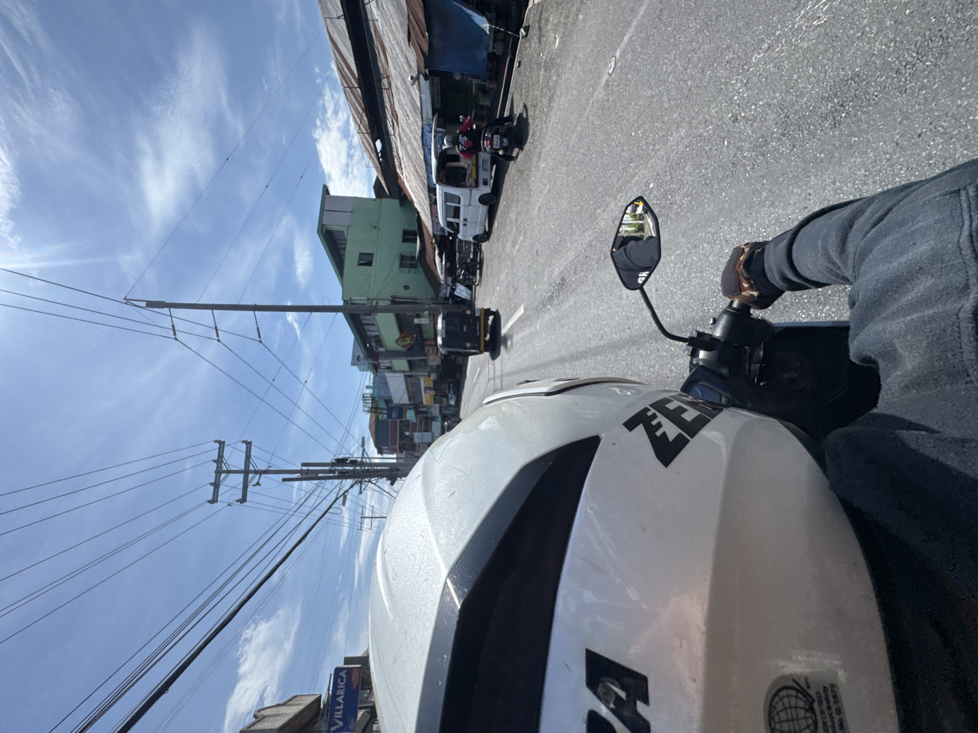 An exterior, eye-level, medium shot shows the perspective of a person riding a motorcycle in Davao City, Philippines. The rider's white helmet, with black accents and a partially visible sticker, is in the foreground. The rider's gloved left hand is on the handlebar. The road ahead is paved and appears to be a commercial street with shops and buildings lining both sides. A white van is parked on the right side of the road, with its rear doors open. A person on a motorcycle is parked behind the van. Further down the street, another motorcycle is seen. Several power lines and utility poles are visible overhead against a bright blue sky with scattered clouds. On the left side of the road, a sign with the word "VILLARICA" is visible. The overall scene depicts a typical daytime street view in a city.