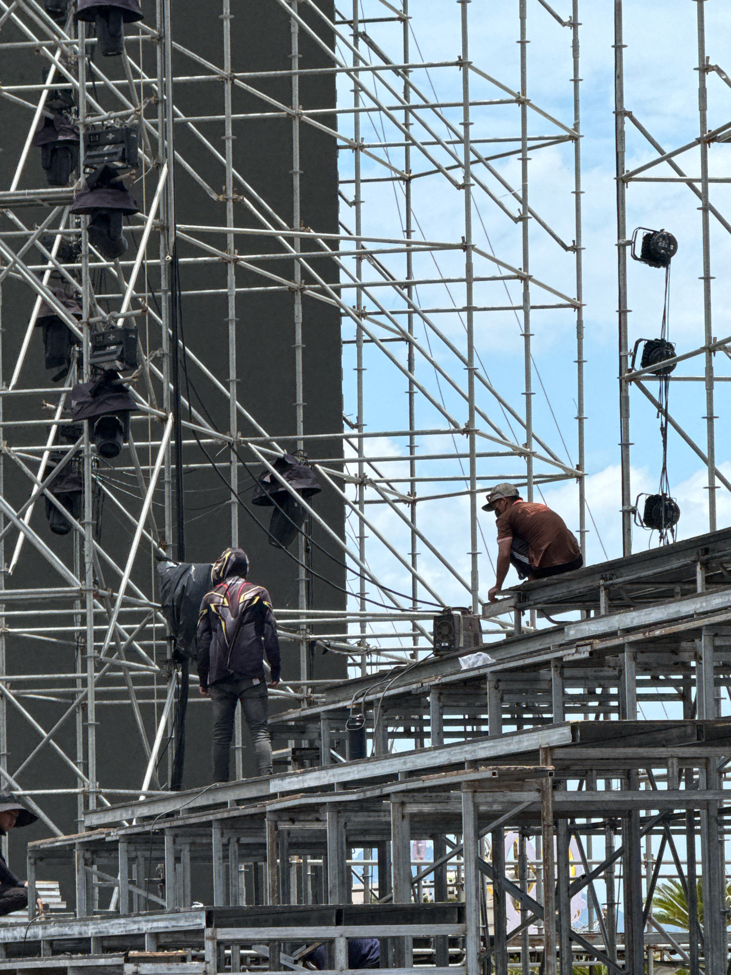 An exterior, eye-level, medium shot captures a large stage under construction in Nha Trang, Vietnam. Several workers, appearing to be of Southeast Asian descent, are visible on the scaffolding. One worker in a brown t-shirt and dark shorts is crouched on a horizontal beam, facing away from the camera, appearing to be working. Another worker, wearing a black jacket with gold and red accents, stands on a higher level of the scaffolding, facing away from the camera. A third worker, in a dark t-shirt and cap, is partially visible in the lower left corner, also facing away. The scaffolding is made of metal, with a large dark screen visible behind it. Several spotlights are attached to the scaffolding. The sky is visible in the background with some clouds. The overall scene depicts the preparatory stages of a large outdoor event, likely a concert or festival, taking place in a coastal city given the palm trees visible in the lower right portion of the frame.