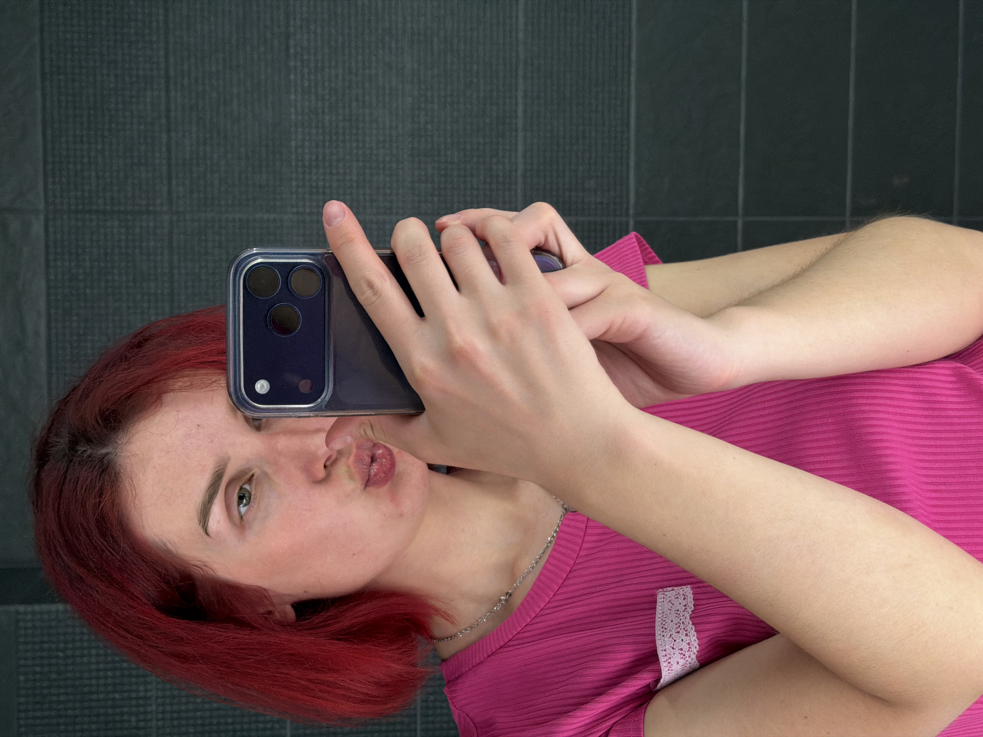 A young woman with red hair is taking a selfie with a smartphone in a room with a gray tiled wall. She is wearing a bright pink shirt and a silver necklace. The phone, encased in a clear case, partially obscures her face. The room appears to be indoors, possibly a bathroom or a similar space with tiled walls. The photograph was taken in the city of Chaikovsky, Russia.