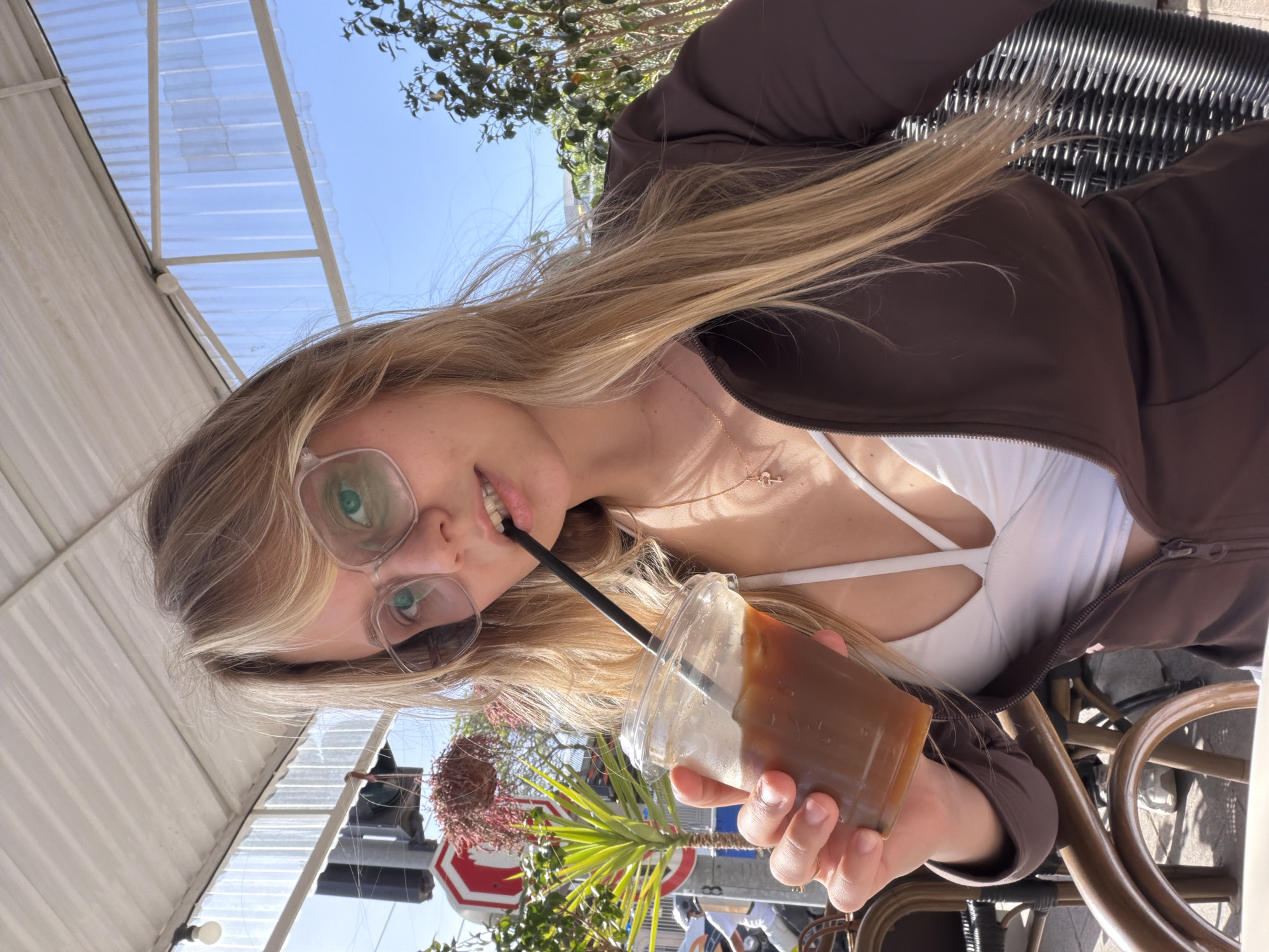 A young woman with blonde hair and glasses is holding a plastic cup with a brown liquid, likely a cold coffee drink, and a black straw, in what appears to be an outdoor cafe setting. She is wearing a brown zip-up jacket and a white top with straps. The location is Bat Yam, Israel, indicated by the visible street signs, including a "STOP" sign, and the surrounding urban environment with plants and buildings. The photo is taken at an angle, showing the woman's face, upper body, and the immediate surroundings of the outdoor seating area, which includes a partially visible rattan chair and a translucent awning. The background shows glimpses of other people and urban scenery.