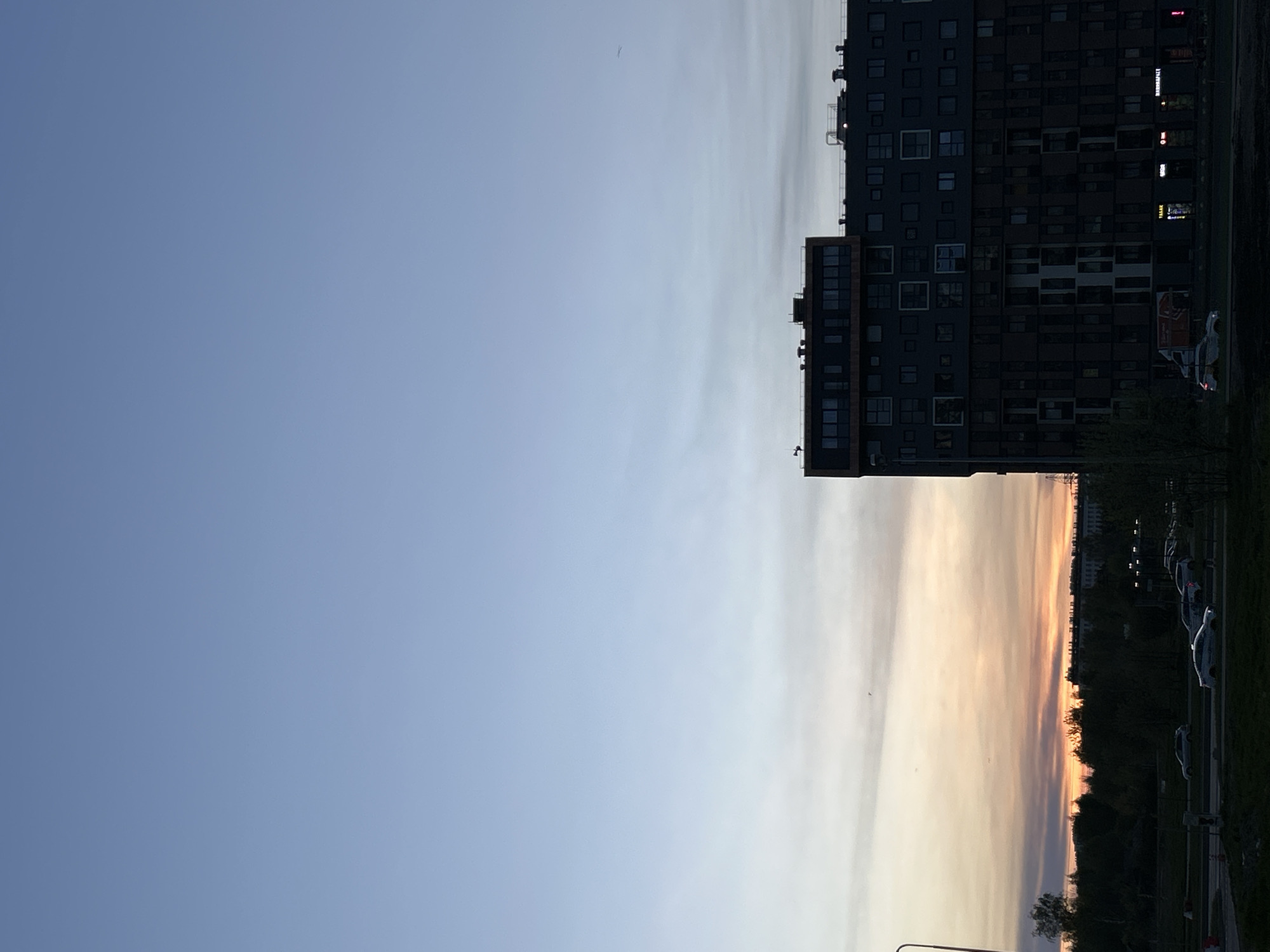The image shows a cityscape at sunset or sunrise. The sky transitions from light blue on the left to a pale orange-yellow on the right. A tall, dark building with many windows is visible on the right. Along the bottom right edge of the image, there is a row of trees and parked cars. The photo appears to have been taken from the city of Saint Petersburg, Russia.