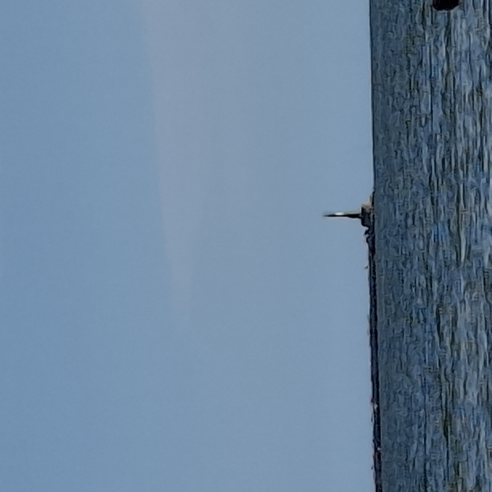 A distant view of a lighthouse and breakwater on the horizon of a body of water, under a clear, light blue sky. The water is a deep blue with visible ripples, suggesting a moderate chop. The lighthouse, dark in color, stands tall on a long, dark breakwater. The breakwater stretches across the image, with its end point featuring the lighthouse. The sky is a uniform, pale blue, devoid of clouds. The overall impression is one of a calm, open seascape.