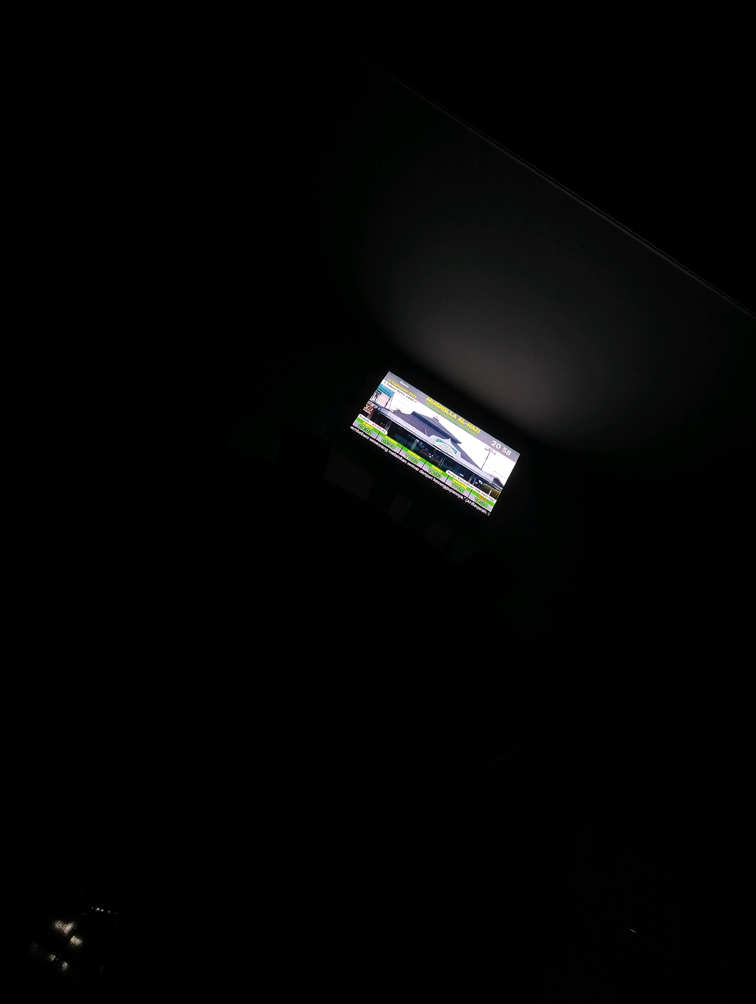 In the dark, a small electronic screen displays an image of a building, likely a mosque, with text in Indonesian. The building is labeled "MUSOLLA AL-HAJJ" and displays prayer times for the day. The screen also shows the date as "Kamis, 16 September 2004" and the time as "20:58". The screen is tilted and appears to be a digital display, possibly a prayer schedule or information board. The surrounding environment is black, suggesting the image was taken at night or in a very dark room, with a spotlight illuminating the screen. No people or actions are visible. This screen may be located in Palangka Raya, Indonesia, as indicated by the Indonesian text.