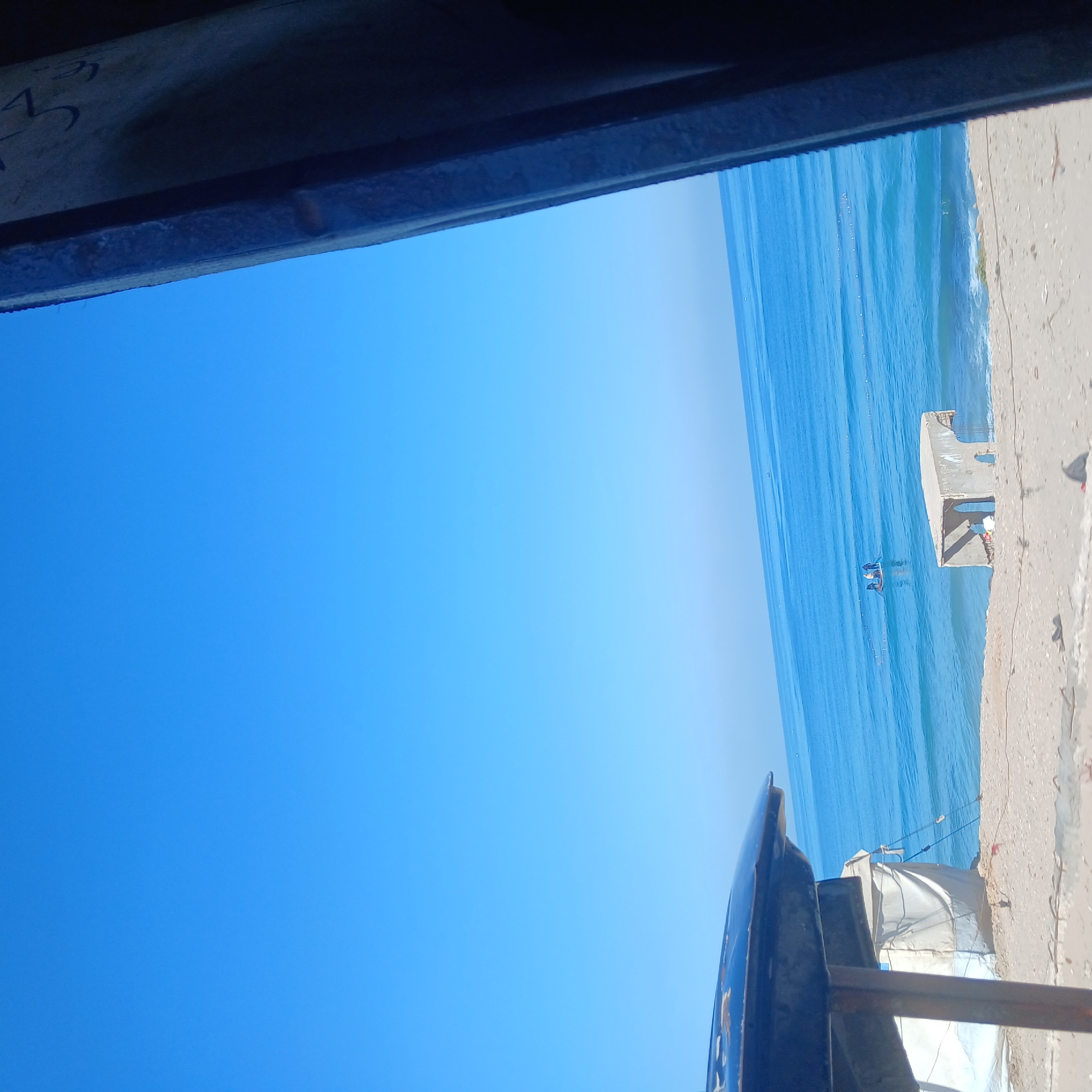 The image depicts a view of the ocean and sky from an elevated position, possibly a building or structure, in an unknown location. The sky is clear and blue, transitioning to a lighter shade near the horizon where it meets the ocean. The ocean is a deep blue with visible wave patterns and what appear to be fishing nets or buoys stretched across the water. A concrete structure is visible along the shoreline, with a small boat nearby. The foreground of the image is dominated by parts of a building or structure, obscuring a portion of the sky and providing a frame for the scene.