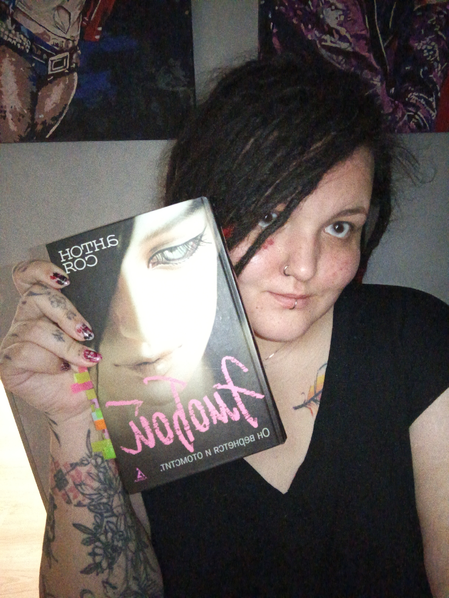 An indoor, head-and-shoulders portrait depicts a young adult female looking directly at the camera while holding a book. The female has dark dreadlocks, a nose ring on her left nostril, and a lip ring centered on her lower lip. Extensive floral tattoos are visible on her left forearm and hand, and a small, stylized feather or flame tattoo is present on her upper chest. She wears a black v-neck shirt. Her fingernails on the visible left hand are painted with a black, red, and white patterned design, accented with neon pink and green.
She holds a book identified as "Nana" by Ai Yazawa, a Japanese manga series. The book's dark cover displays a stylized pale female face, specifically the character Nana Osaki, with distinctive eye makeup. The title "NANA" is printed at the top (appearing as mirrored "HOTHG ROO"), and the author "AI YAZAWA" is below (appearing as mirrored "WODOWOГ"), both in pink text. Additional mirrored text, "INTOМОТО N ЯОТS", is present at the bottom of the cover.
The background consists of a light grey wall. Two framed art pieces are visible: the left shows the lower torso of an athletic figure in a belt and brief-like garment, while the right displays an abstract design predominantly in purple and dark tones. The lighting suggests an interior residential setting. No specific landmarks or unique architectural features are present to pinpoint an exact location within Chaikovsky, Russia.