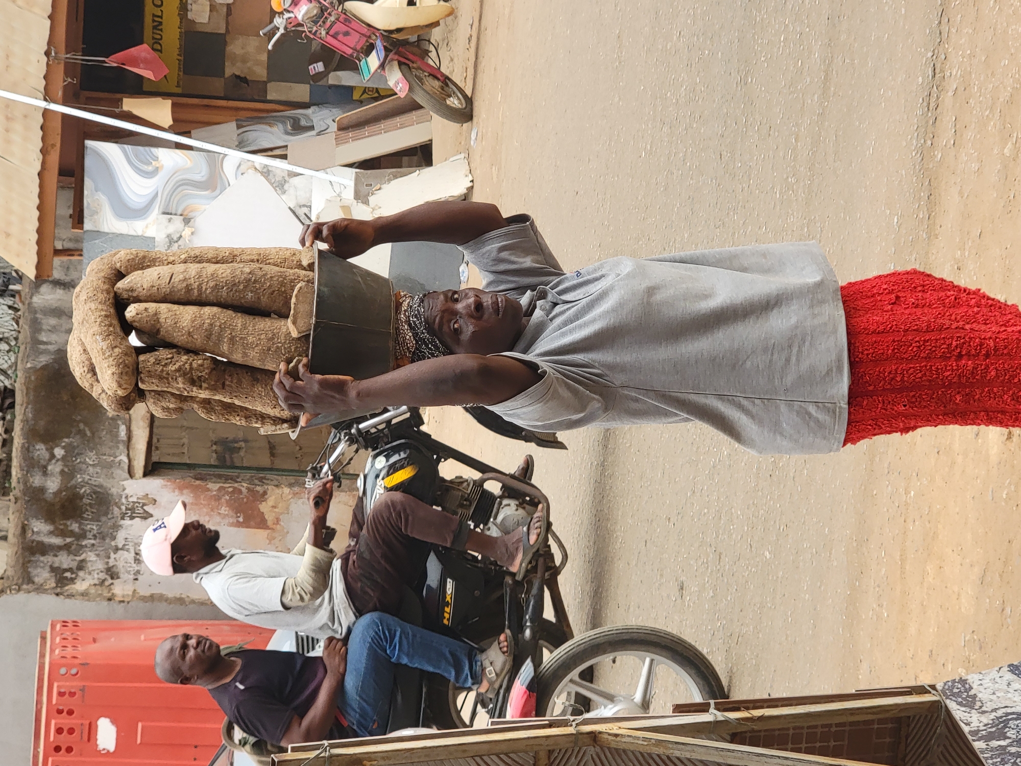 In Maiburiji, Nigeria, an adult woman wearing a gray shirt, red pants, and a patterned headscarf carries a large collection of yams in a black container on her head. She looks toward the viewer. Behind her, a motorcycle is connected to a small carriage carrying two men. One man wears a white shirt and pink baseball cap, while the other wears a dark shirt and blue jeans. The setting appears to be a city street, with buildings and other vehicles visible in the background.