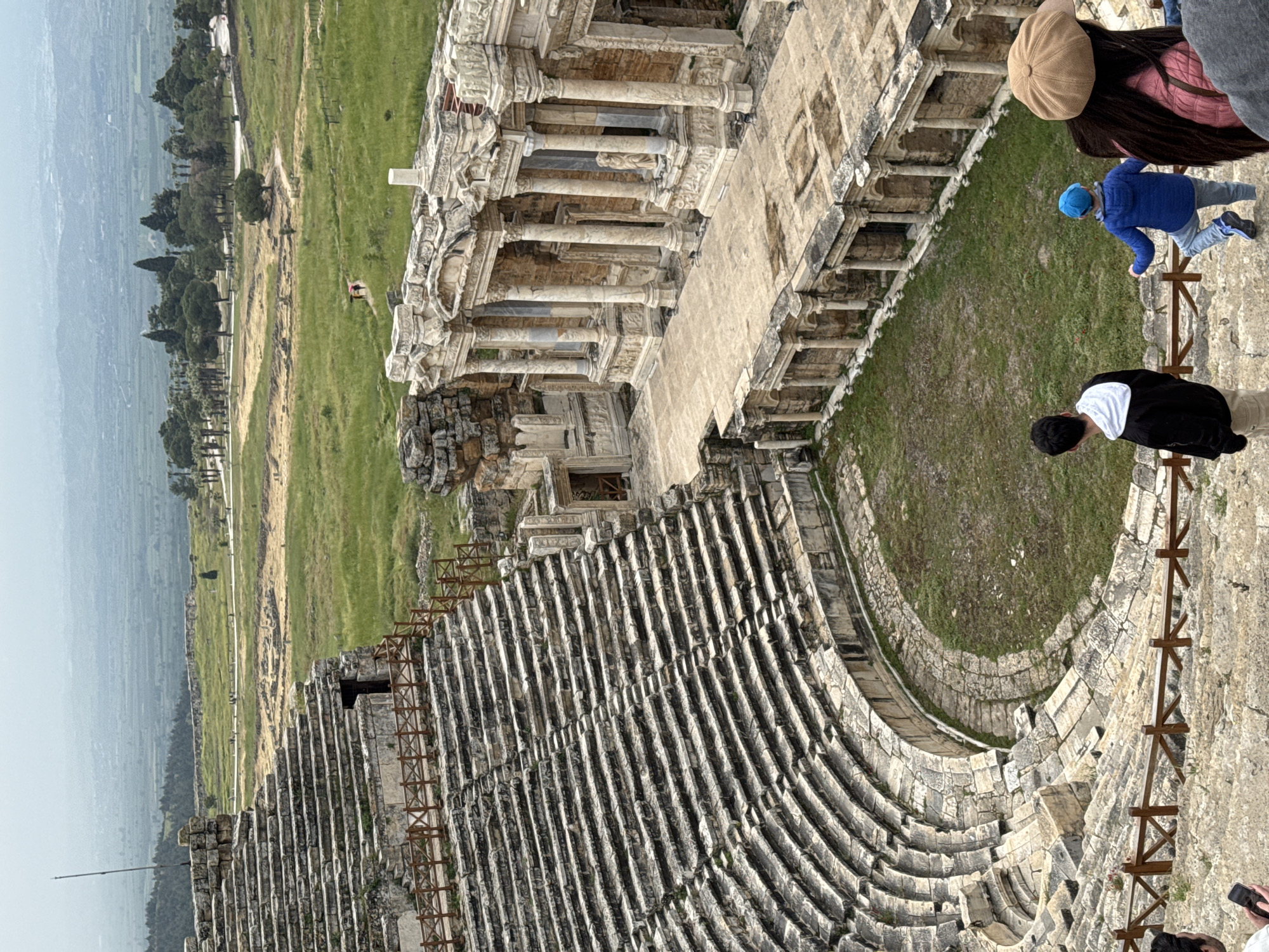The image shows the ancient theater in Pamukkale, Turkey. The theater is a well-preserved Roman structure with tiers of stone seating arranged in a semi-circular shape. Some people stand at the top and bottom of the theater, taking in the view. The stage area is partially intact, with ornate columns and architectural details visible. Beyond the theater lies a vast landscape of green fields and distant mountains, adding to the historical setting. A temporary wooden barricade is in place along the edge of the seating area.