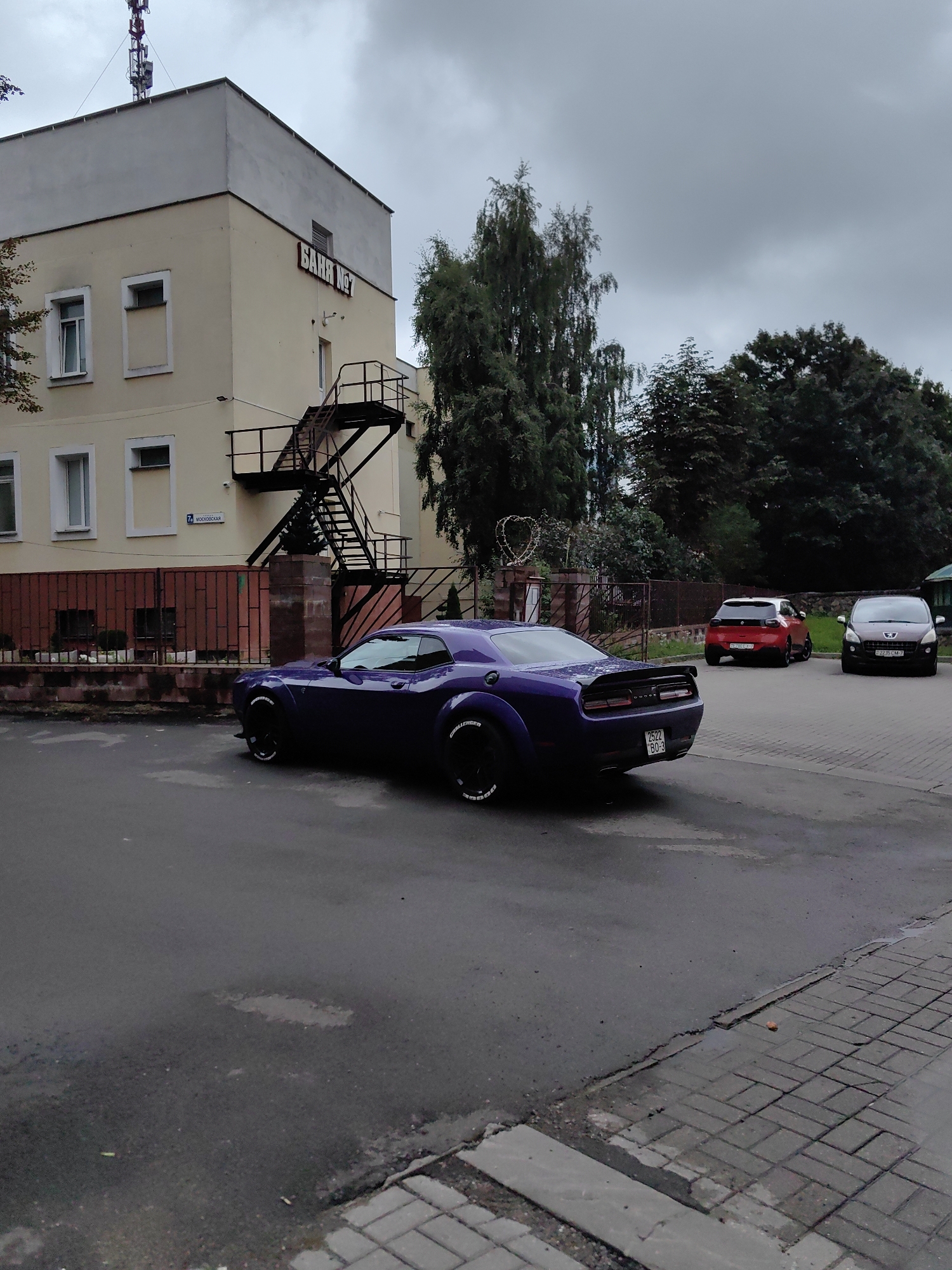 The image depicts an outdoor scene on a cloudy day, likely in Minsk, Belarus, given the car license plates. In the foreground, a dark purple Dodge Challenger is parked on a wet asphalt road. The car has custom white lettering on its tires that reads "CHALLENGER". A portion of a paved pedestrian walkway is visible in the lower right corner.
Behind the car, a light yellow building with a light grey upper section stands. The building has several windows and an external metal fire escape. A sign on the building reads "БАНЯ №7" (Banya No. 7, which translates to "Bathhouse No. 7"). A street sign with "75 МОСКОВСКАЯ" (75 Moskovskaya) is also visible near the building. A metal fence with a decorative heart-shaped wire sculpture is situated in front of the building.
In the background, several trees are present, along with two other parked cars: a red hatchback on the left and a grey hatchback on the right. The red car's license plate appears to be a Belarusian plate, and the grey car's plate is also likely Belarusian. The overall impression is a street scene with residential or commercial buildings and parked vehicles.