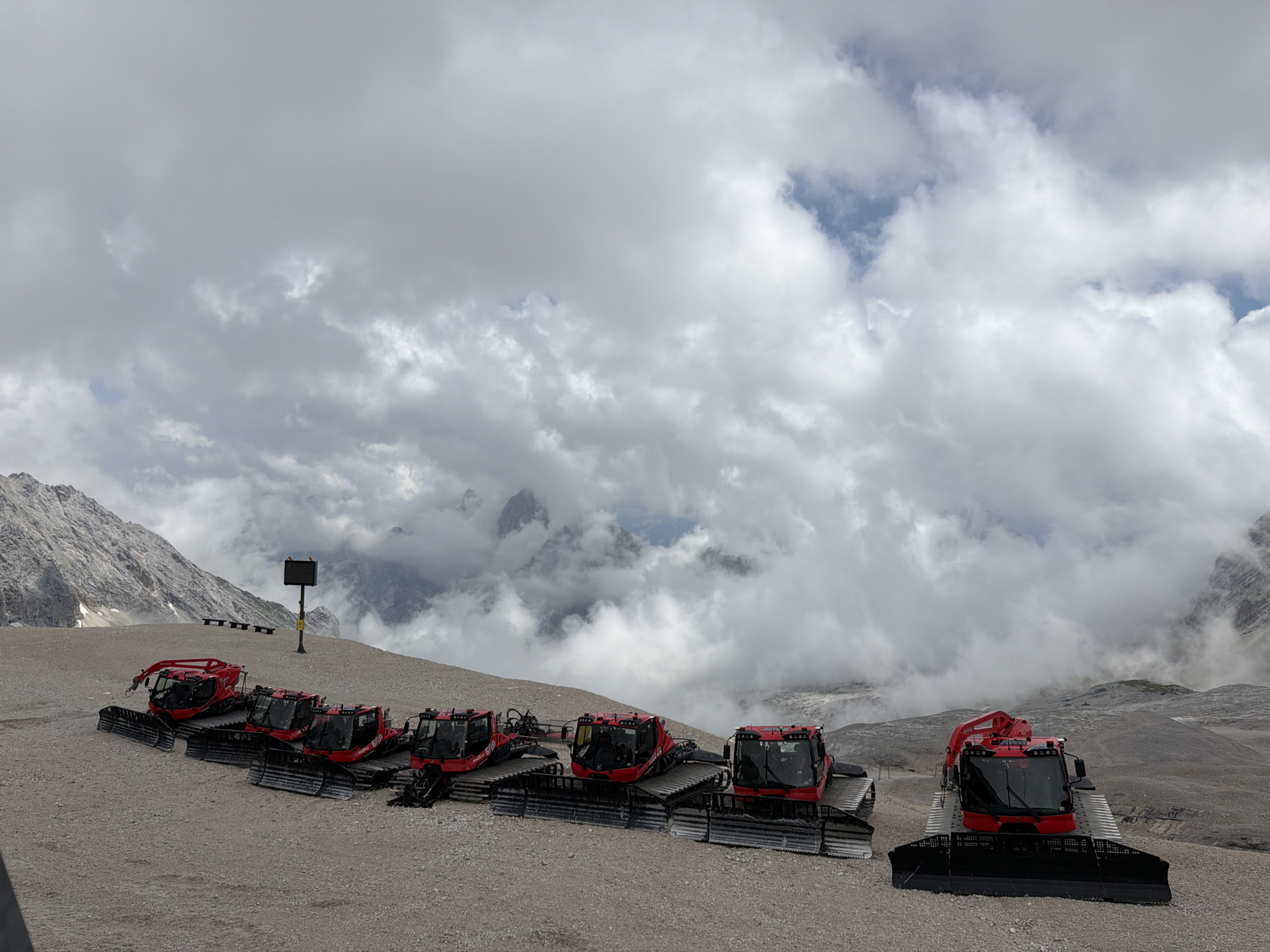 The image depicts a line of red snow groomers parked on a gravelly surface, likely in a mountainous region. The snow groomers are the primary subjects, all facing generally towards the right of the frame. They have large treads and a plow-like attachment at the front. The background is dominated by a cloudy sky with patches of blue visible. In the distance, mountains are visible, with some shrouded in mist. A single signpost with a black panel is also present on the left side of the image. The scene appears to be outdoors in a high-altitude environment, possibly a ski resort. While the exact location in Garmisch-Partenkirchen, Germany is not explicitly stated in the image, the presence of snow groomers and mountainous terrain suggests it could be a ski area in the vicinity of the Zugspitze, Germany's highest peak, which is located near Garmisch-Partenkirchen. There are no people visible in the image.