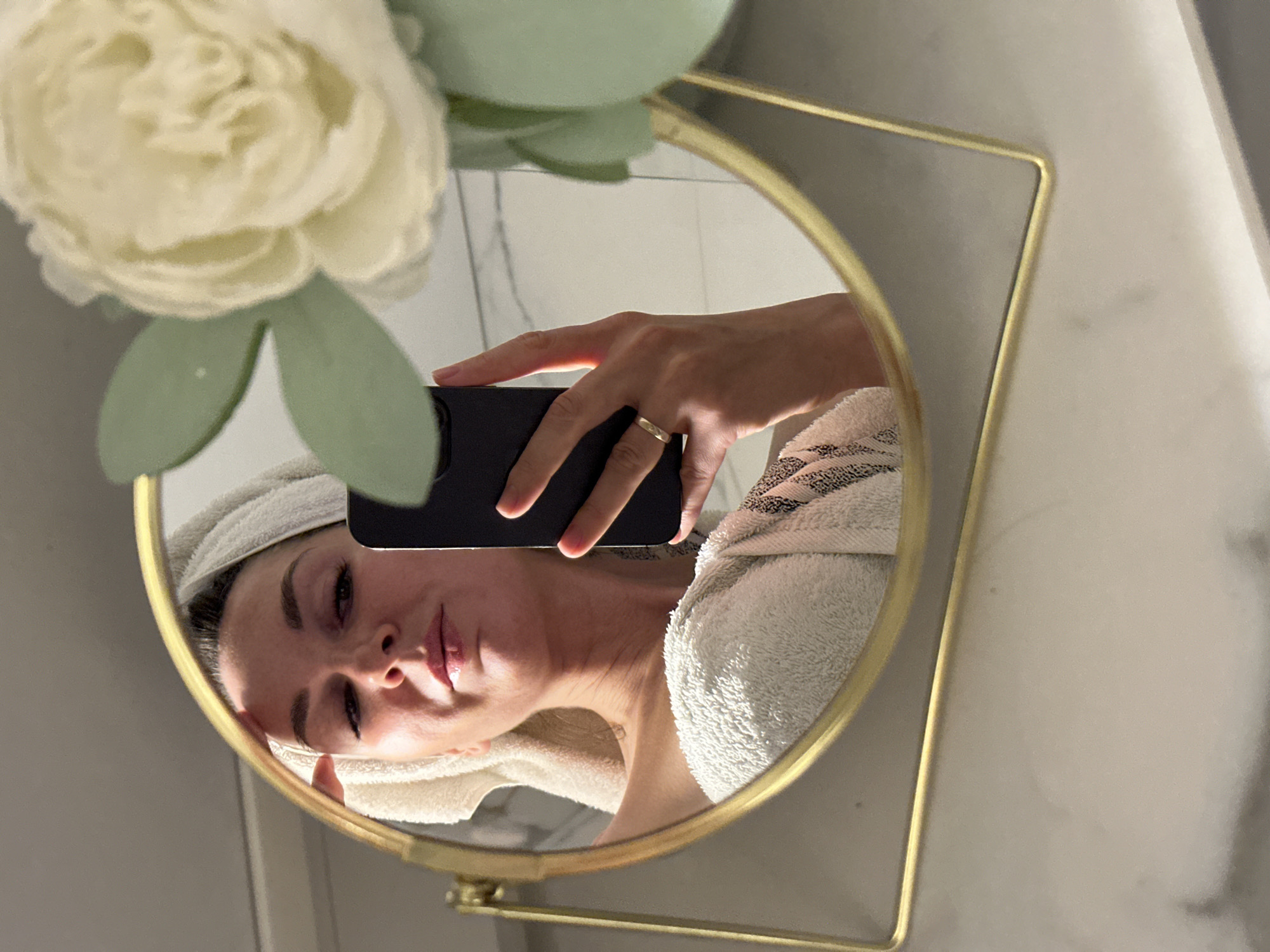 A woman is taking a mirror selfie in what appears to be a bathroom. She is wearing a white towel wrapped around her head and another towel draped over her shoulders. She is holding a smartphone in her right hand, and her left hand is touching her forehead. A decorative artificial flower is visible in the foreground. The reflection shows a woman with a towel on her head, partially wrapped in a towel, holding a phone and touching her forehead. The background reflection appears to be a tiled wall. There is no indication of the location being in Chaikovsky, Russia.