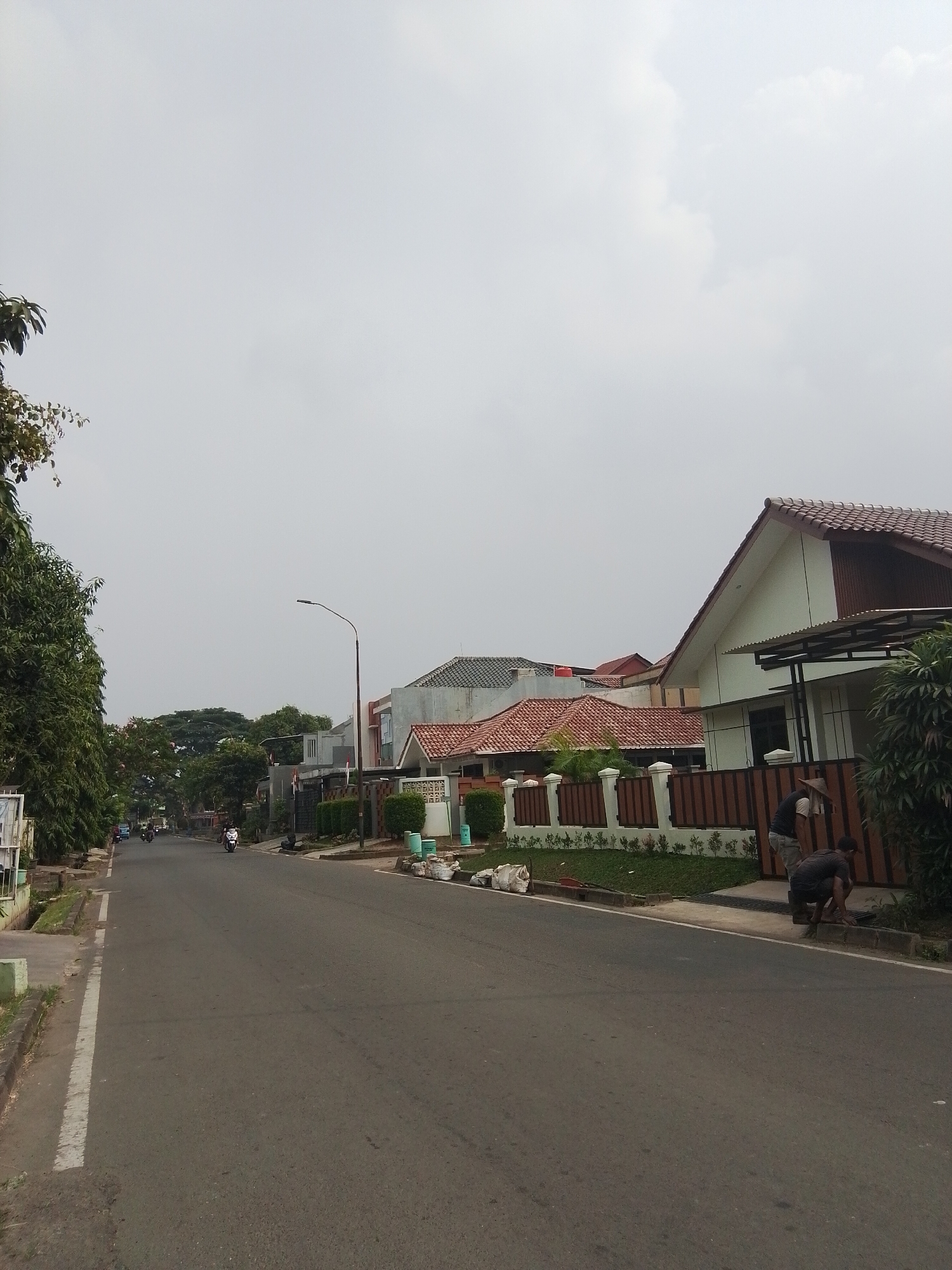 This is a vertical, eye-level shot of a residential street in Bencongan, Indonesia, on an overcast day. The asphalt road occupies the lower half of the frame, with a white line marking the edge of the lane. On the left, a sidewalk with a small drainage ditch runs parallel to the road, bordered by trees and a section of a building. Several motorcycles are visible on the road, some in motion. The right side of the street features a row of houses with tiled roofs, fences, and manicured lawns. Two men are present in the foreground on the right, with one crouching and the other standing. They appear to be engaged in some form of maintenance work near the roadside. The background is dominated by a pale sky with scattered clouds, and a line of trees is visible in the distance.