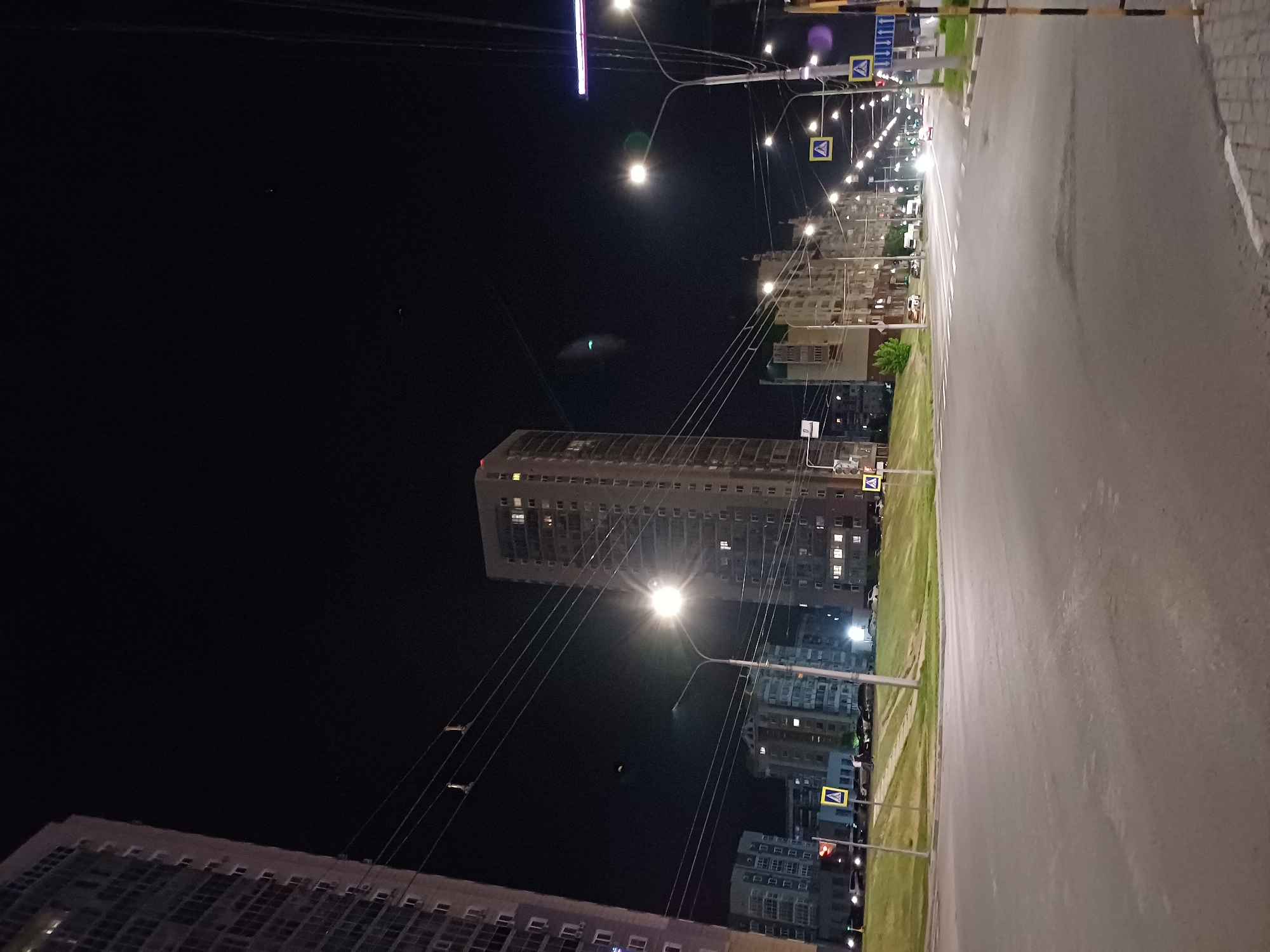 An eye-level, vertical, night time shot captures a street scene in a city. Tall residential buildings dominate the background, their windows illuminated, suggesting occupancy. A streetlamp with a bright, central light source illuminates the foreground, casting a glow on the wet asphalt road. Multiple pedestrian crossing signs, marked with a yellow triangle and a pedestrian icon, are visible along the roadside. A few vehicles are present, some parked and others in motion, their headlights contributing to the nighttime illumination. The sky is dark and clear, with no visible celestial bodies. The overall environment appears to be urban, with sidewalks and grassy areas adjacent to the road.