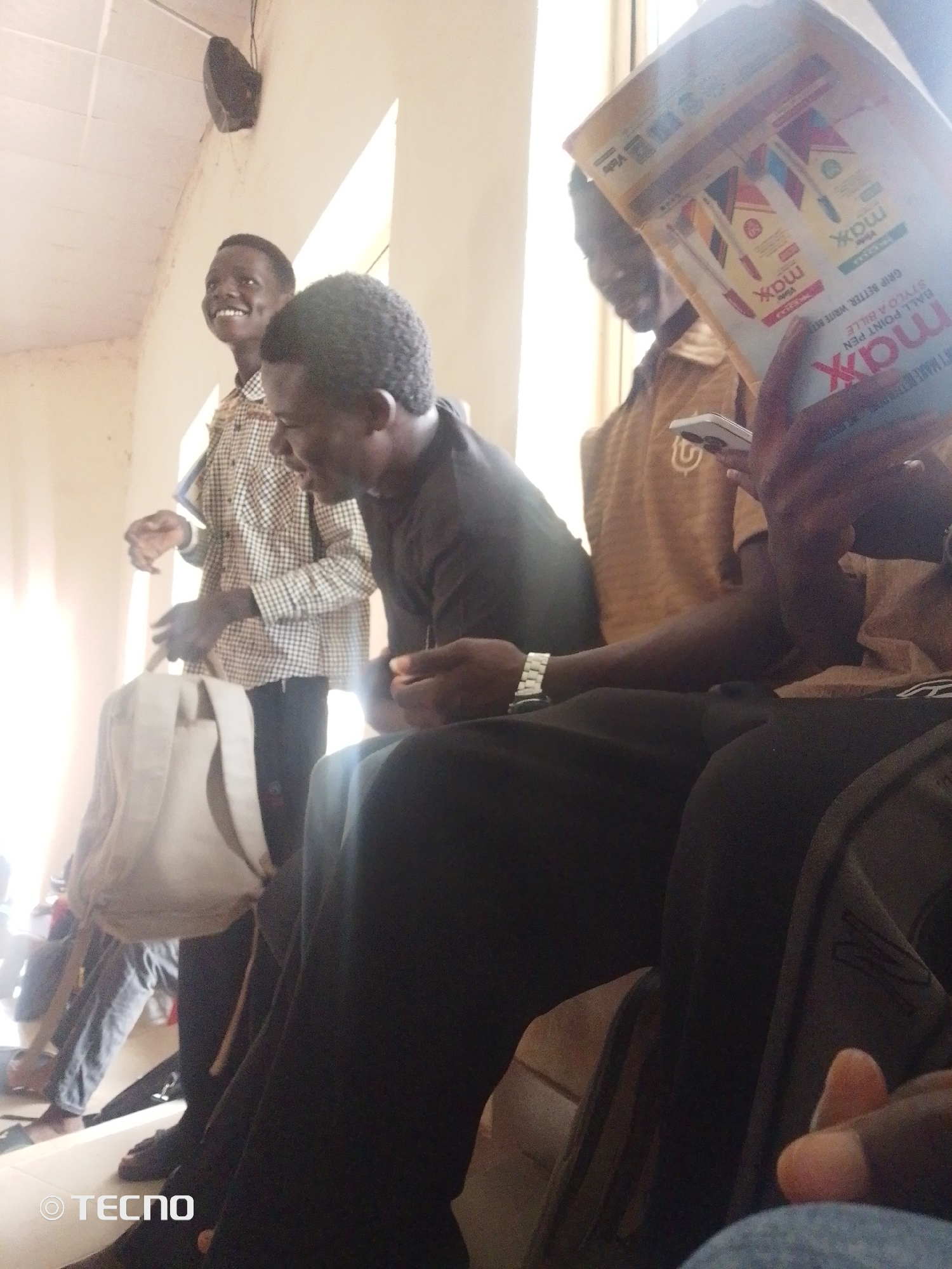 A group of young men are gathered in what appears to be a classroom or community hall in Gumel, Nigeria. The lighting suggests it is daytime, with natural light streaming in from a window or opening. In the foreground, a young man in a black t-shirt is seated, looking down and to the right. To his left, another young man wearing a checkered shirt is standing and smiling at the camera, with a white backpack slung over his shoulder. Further back and to the right, two other young men are visible. One is wearing a brown shirt with a "U" emblem and is holding a mobile phone. Another person is partially obscured by the group, holding a magazine or pamphlet, which displays advertisements for "Max" brand pens. The overall atmosphere seems casual and convivial.