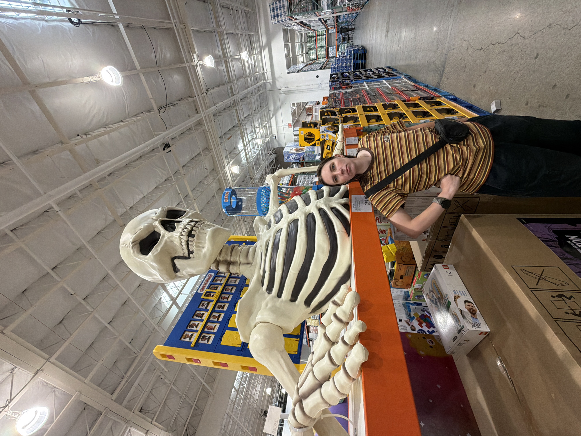 A person stands next to a large skeleton decoration inside a Costco store in Beaverton, Oregon, United States of America. The person is wearing a striped t-shirt and dark pants, with a black bag slung across their chest. The skeleton is a prominent display, with its head and upper body visible. In the background, shelves are stocked with various merchandise, and aisles of the warehouse store are visible. The ceiling is high with exposed beams and industrial lighting.