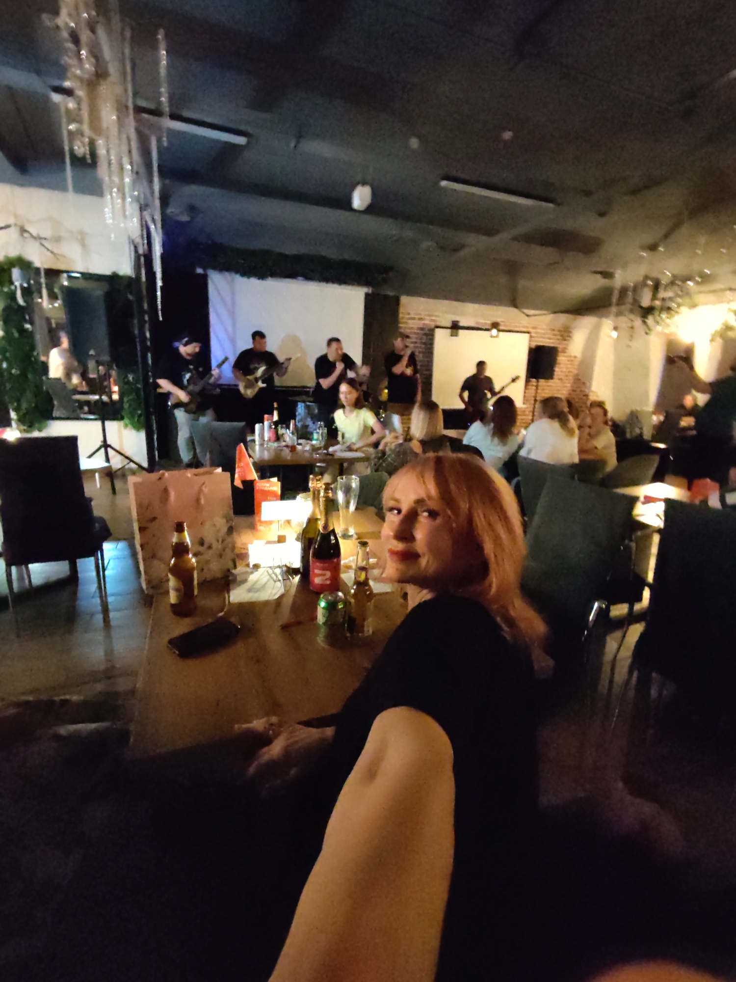 A woman with blonde hair and red lipstick smiles at the camera while seated at a table in a dimly lit restaurant or bar, likely in Chaikovsky, Russia. She is wearing a black top and is partially visible in the foreground. Behind her, a band is performing on a stage with a white backdrop. Several other patrons are seated at tables, some in the background, and some closer to the band. On the table in front of the woman, there are several bottles, a can, and a smartphone. The environment appears to be indoors, with exposed brick walls and decorative lighting.