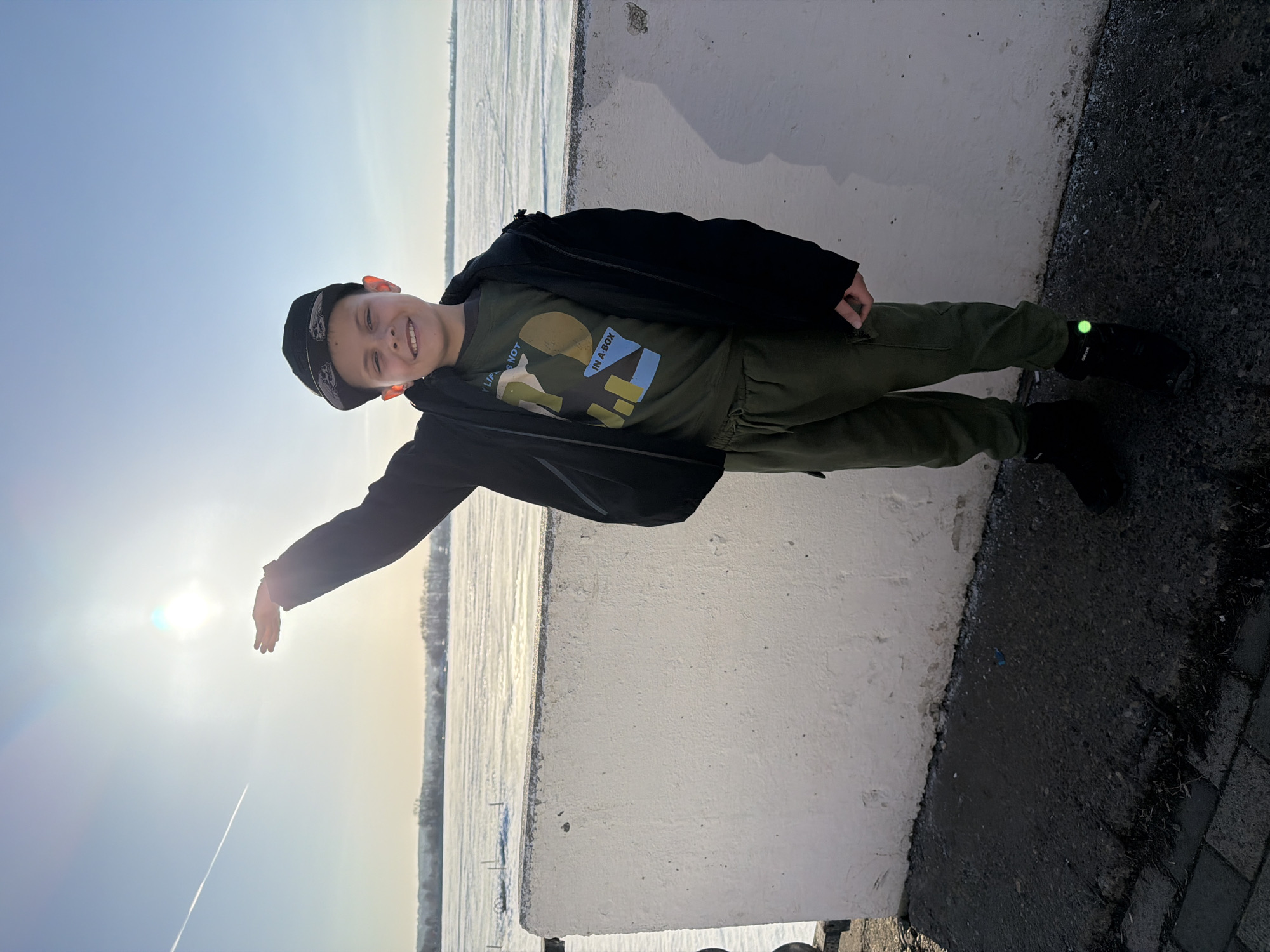 A young boy stands next to a concrete barrier, reaching his arm towards the sun. He is wearing a dark jacket, green shirt, green pants, and black shoes. His footwear has a green reflective detail. He wears a cap on his head. Behind him is a body of water, likely a portion of the Votkinsk Reservoir, with a layer of ice visible. The location is likely near a waterfront area in Votkinsk, Russia.