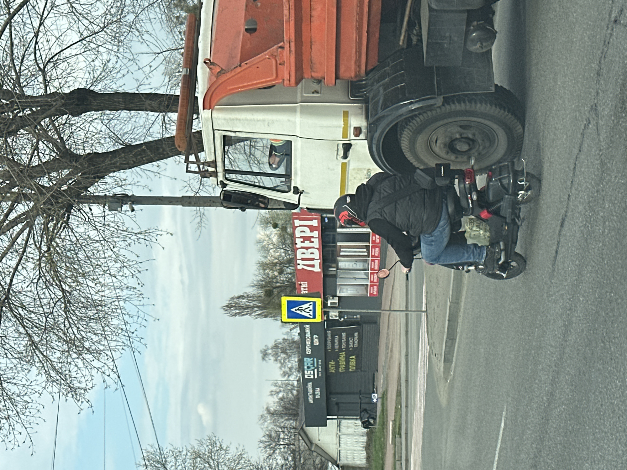 The image captures a street scene, potentially in the city of Kyiv, Ukraine, with text signs displayed in Ukrainian. A large white and orange truck is partially visible on the right. A man on a moped is positioned near the truck's rear. Behind him, signs indicate "ДВЕРІ" (Doors) and a pedestrian crossing. Also, there are signs related to "АНТИГРАВІЙНА ПЛІВКА" (Anti-gravel film) and other services. The trees in the image appear bare, suggesting a cold season.