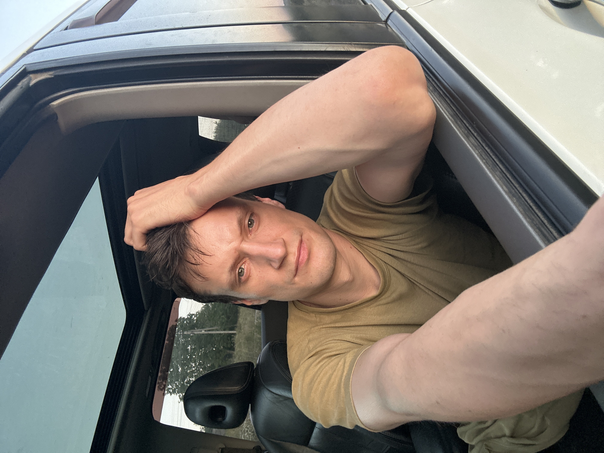 A medium shot captures a Caucasian male with short brown hair, his right hand resting on the top of his head. He is seated in the driver's side of a white SUV, with his left arm resting on the open car door. The man is wearing a short-sleeved olive green t-shirt. The interior of the car features black leather seats and a sunroof. Outside the car, a blurred background suggests a rural setting with trees and utility poles visible. The lighting indicates it is late afternoon or early evening. The image is an outdoor selfie taken from inside a vehicle. The location is Makiv, Ukraine.