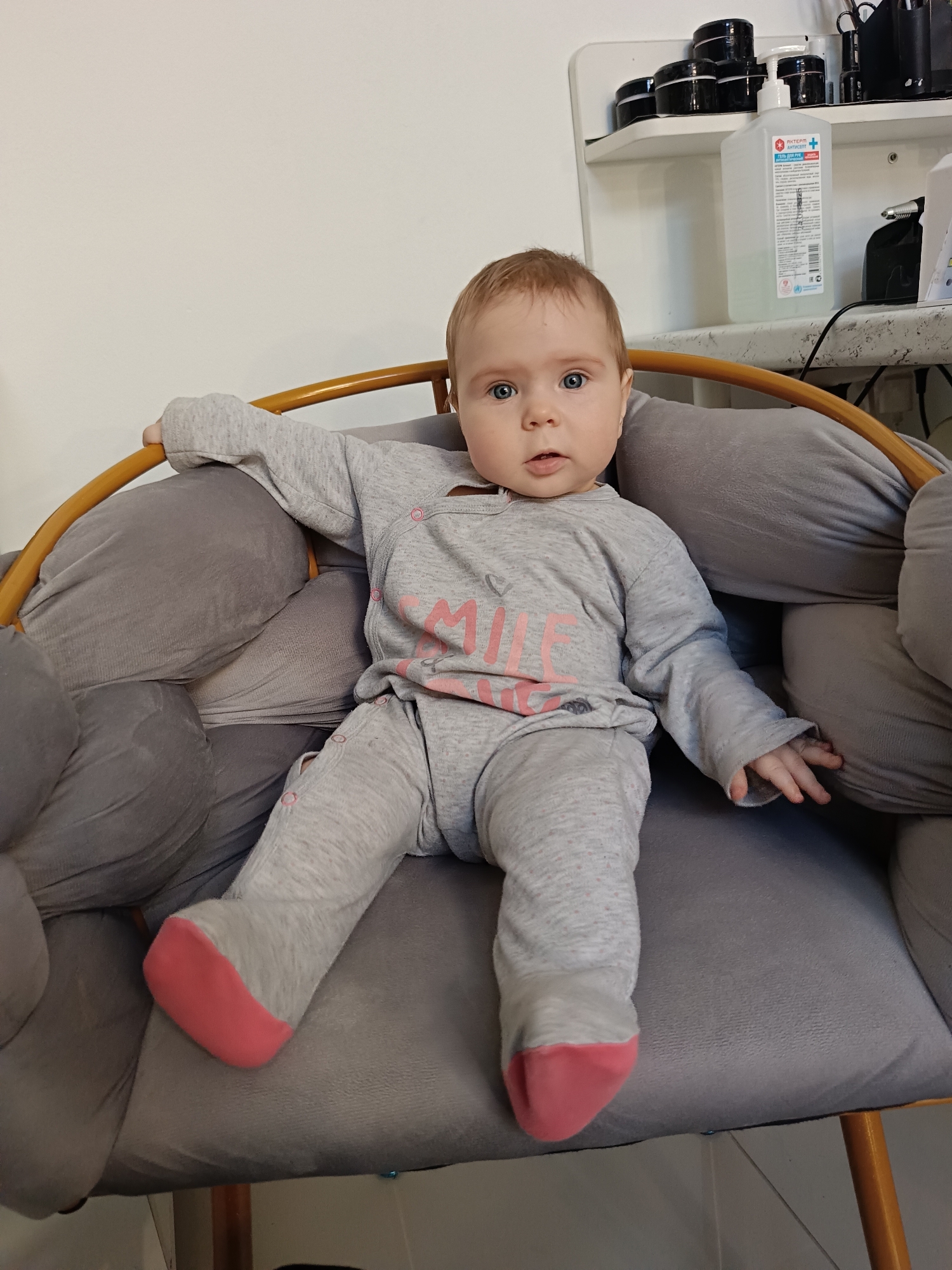 A baby with red hair and blue eyes is sitting in a grey chair with a wooden frame. The baby is wearing a grey outfit with pink accents and the word "SMILE" on the front. The baby is looking directly at the camera with a slightly open mouth. Behind the chair, there is a white shelf with various containers and a bottle of hand sanitizer. The scene is indoors. It is located in the city of Chaikovsky, Russia.