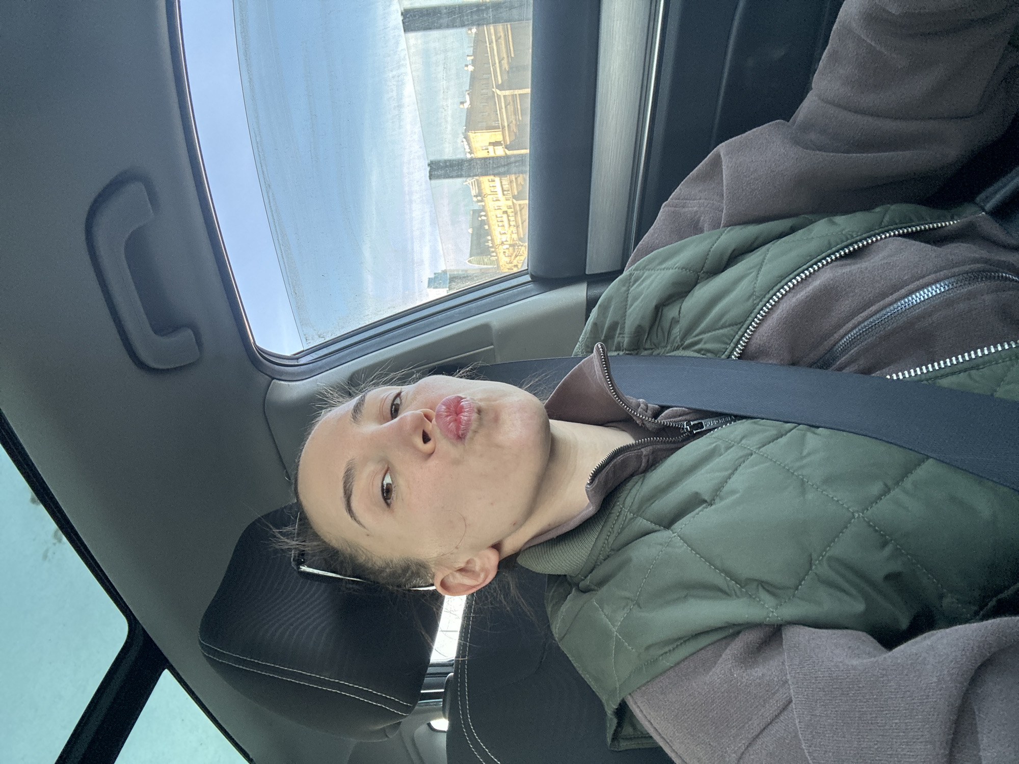 This media file captures a young woman, likely in her late teens or early twenties, seated in the passenger seat of a car. She is looking directly at the camera with a playful "pouty" or "kissing" expression, her head slightly tilted. She has light skin, dark eyebrows, and brown eyes, with her hair appearing to be pulled back.
She is dressed in layers, wearing a dark brown or mauve long-sleeved top or hoodie, over which she has a dark green quilted vest. A black seatbelt is fastened across her chest and shoulder. The interior of the car is visible, featuring a black headrest with white stitching behind her and an overhead grab handle to the left.
Through the car's windows, an urban environment, specified as Moscow, Russia, is seen under a clear blue sky. A sunroof above shows the bright, cloudless sky, indicating a daytime setting with fair weather. Through the side window to her right, several light-colored, multi-story buildings with numerous rectangular windows are visible, suggestive of city architecture. There appears to be some dust or reflections on the window glass. The overall atmosphere conveyed is casual and lighthearted. There is no visible text in the image.